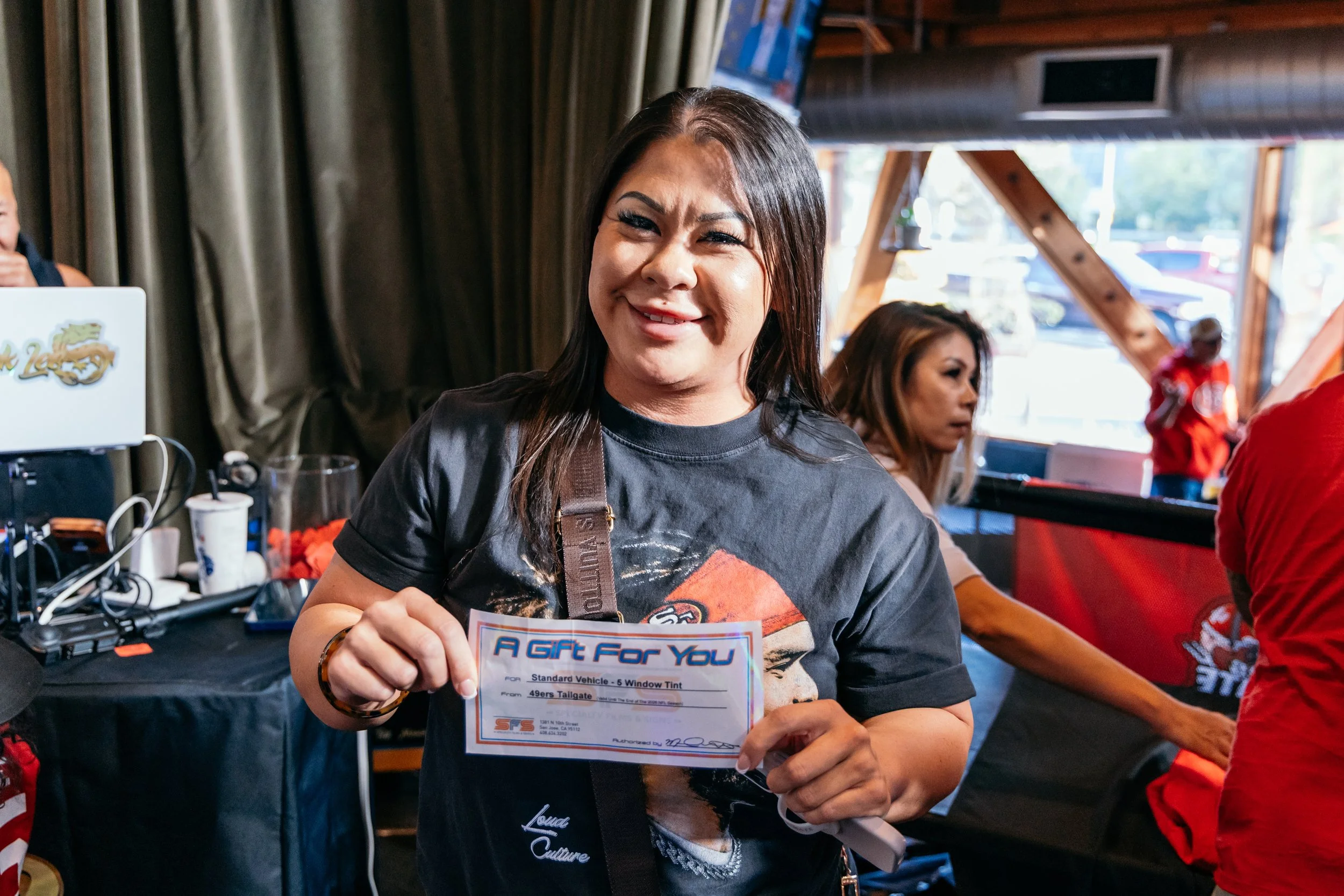 Woman smiling and holding a flyer that says 'A Gift For You' at an indoor event with others in the background, including a person in a red shirt.