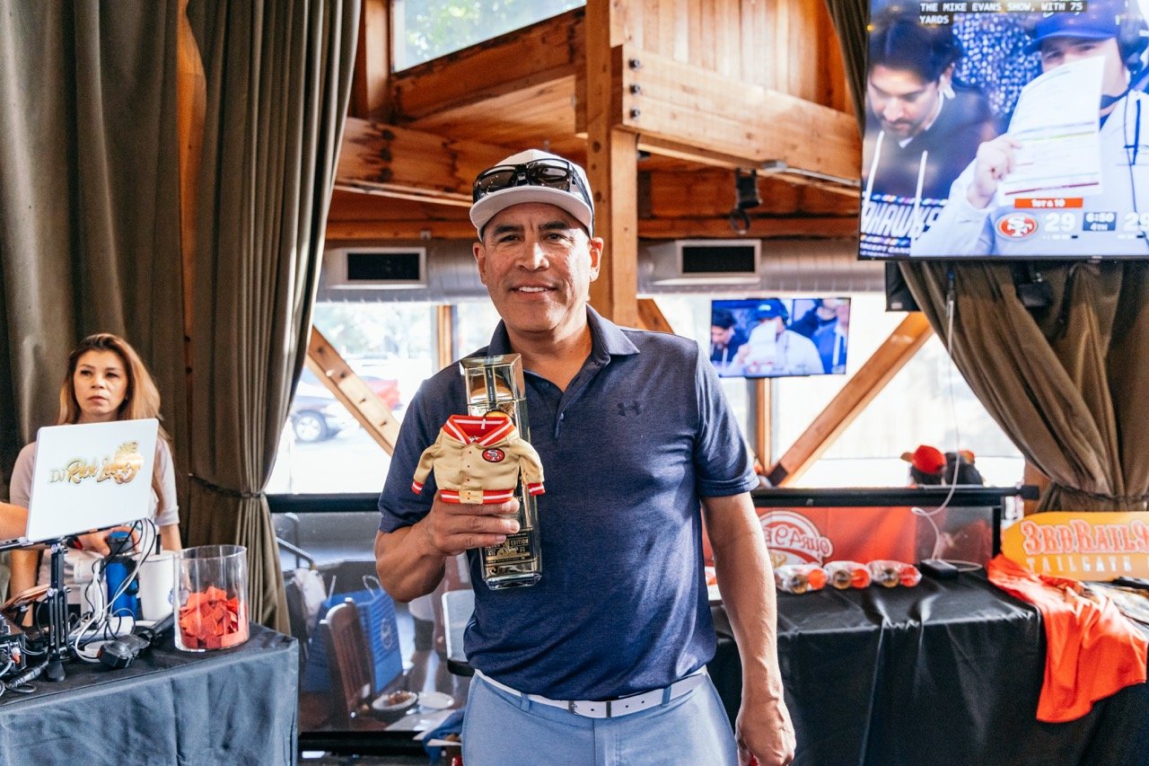 A man is holding a bottle of liquor with a small foam New England Patriots hockey jersey attached to it, smiling at the camera in a festive setting. There are multiple TVs in the background showing a sports game, and a woman is sitting at a table wit