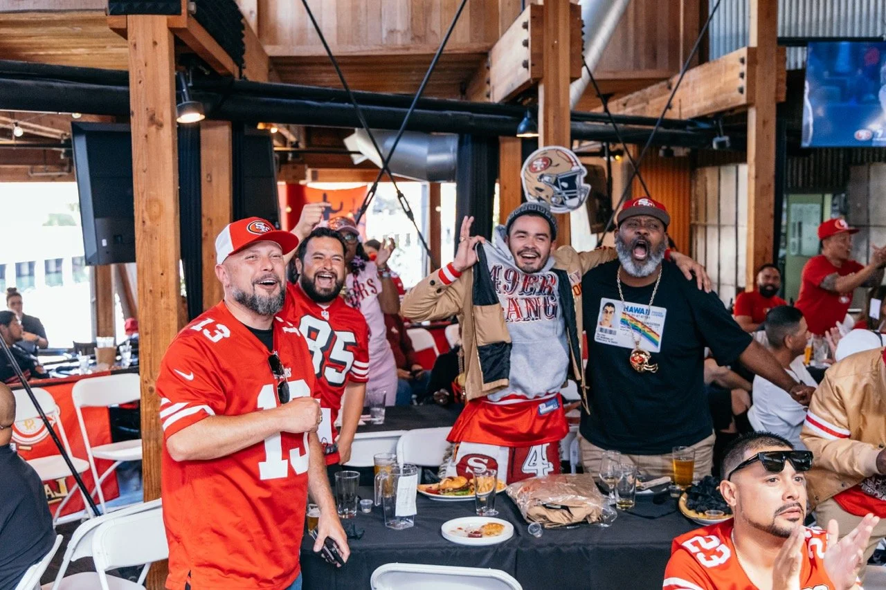 A group of people wearing San Francisco 49ers jerseys and hats, celebrating and smiling at a sports event or party indoors.