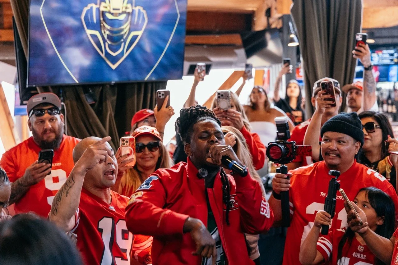 A group of people, mostly wearing red sports jerseys, gathered in a lively indoor setting. They are taking photos and videos with their smartphones, some smiling and others singing or talking. A man in the center, with dreadlocks, is singing into a m