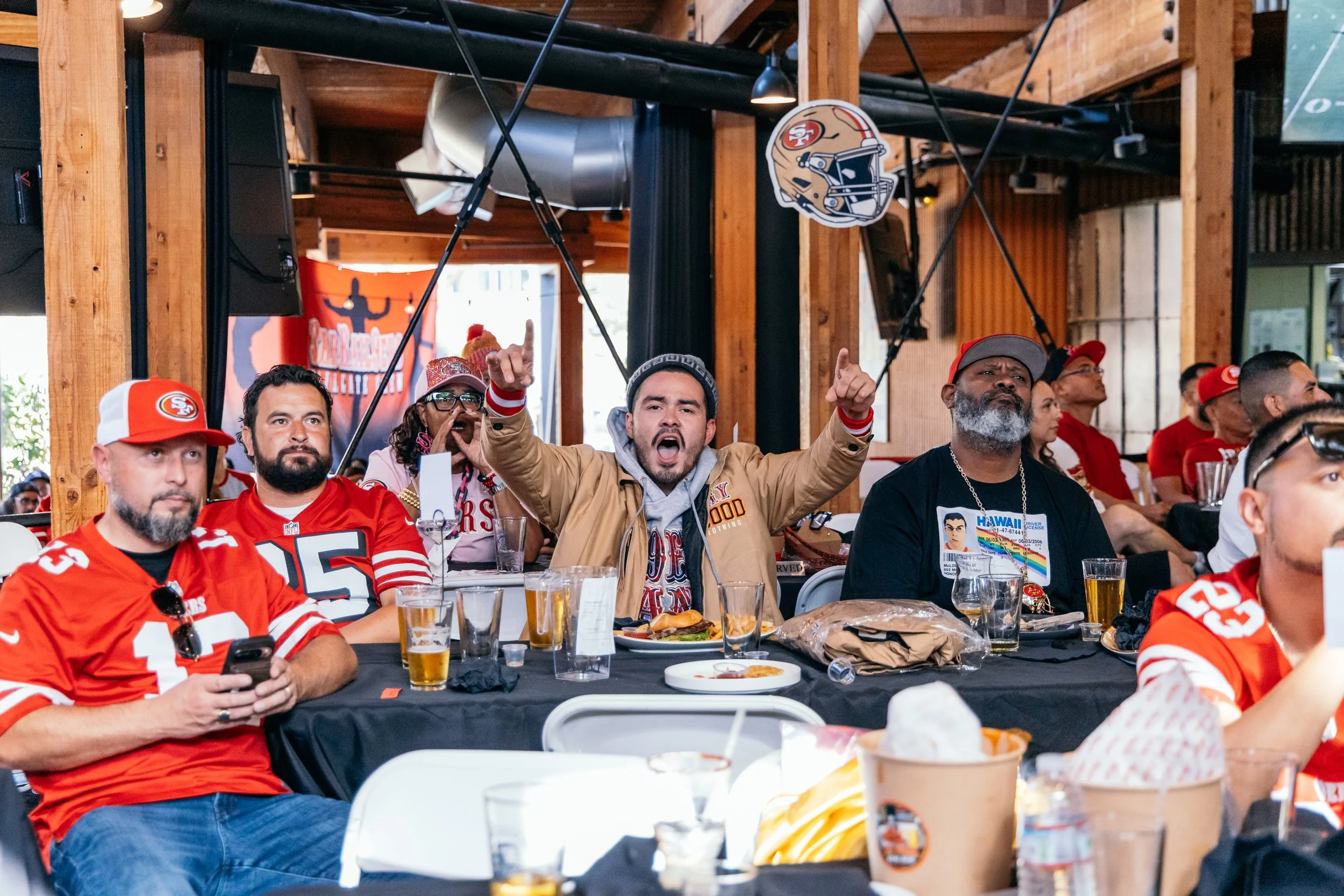A group of diverse people, mostly wearing San Francisco 49ers football jerseys, sitting at a long table in a wooden-furnished room, watching and reacting to a sports event on a screen. Some are cheering, raising their hands, or looking focused. There