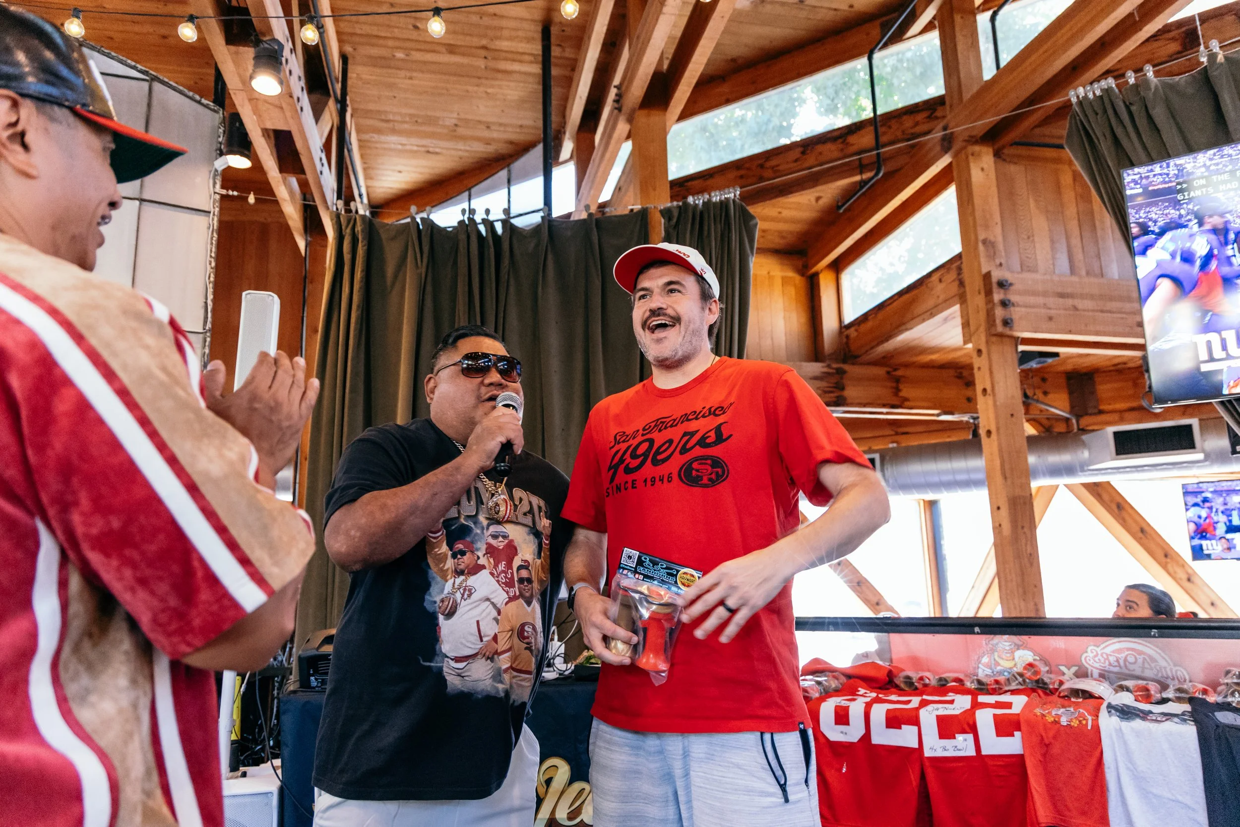 A group of men wearing San Francisco 49ers apparel and accessories are gathered indoors, celebrating or speaking during an event. One man is holding a microphone, and another man is smiling, holding what appears to be a product or prize. The backgrou