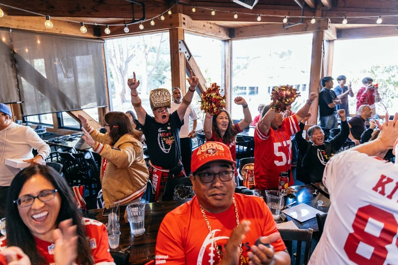 A group of people in a restaurant celebrating, wearing San Francisco 49ers football jerseys and apparel, cheering, clapping, and raising their arms in excitement.