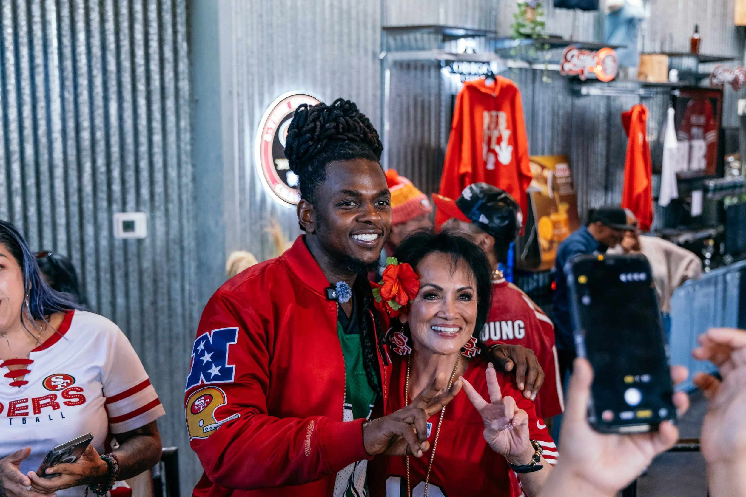 Group of people at a celebration party wearing San Francisco 49ers attire, smiling and taking photos, with a woman wearing a red jacket and a woman wearing a white shirt with red accents in the foreground.