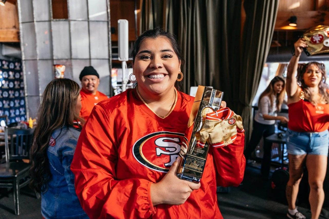 A woman in a red San Francisco 49ers shirt holding a trophy and a small bag, smiling at the camera in a crowded indoor setting with other women in the background.