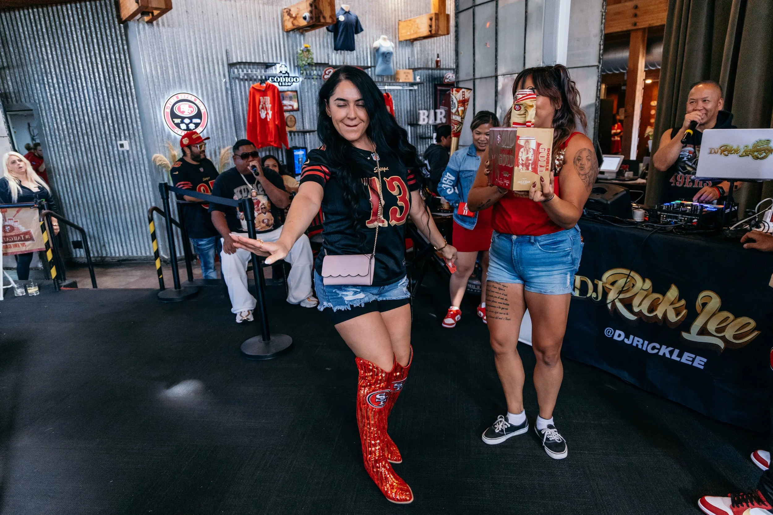 Two women celebrating at a San Francisco 49ers themed event, one wearing 49ers-themed red sequined boots and the other holding a gift box, surrounded by people, with a DJ stage in the background.