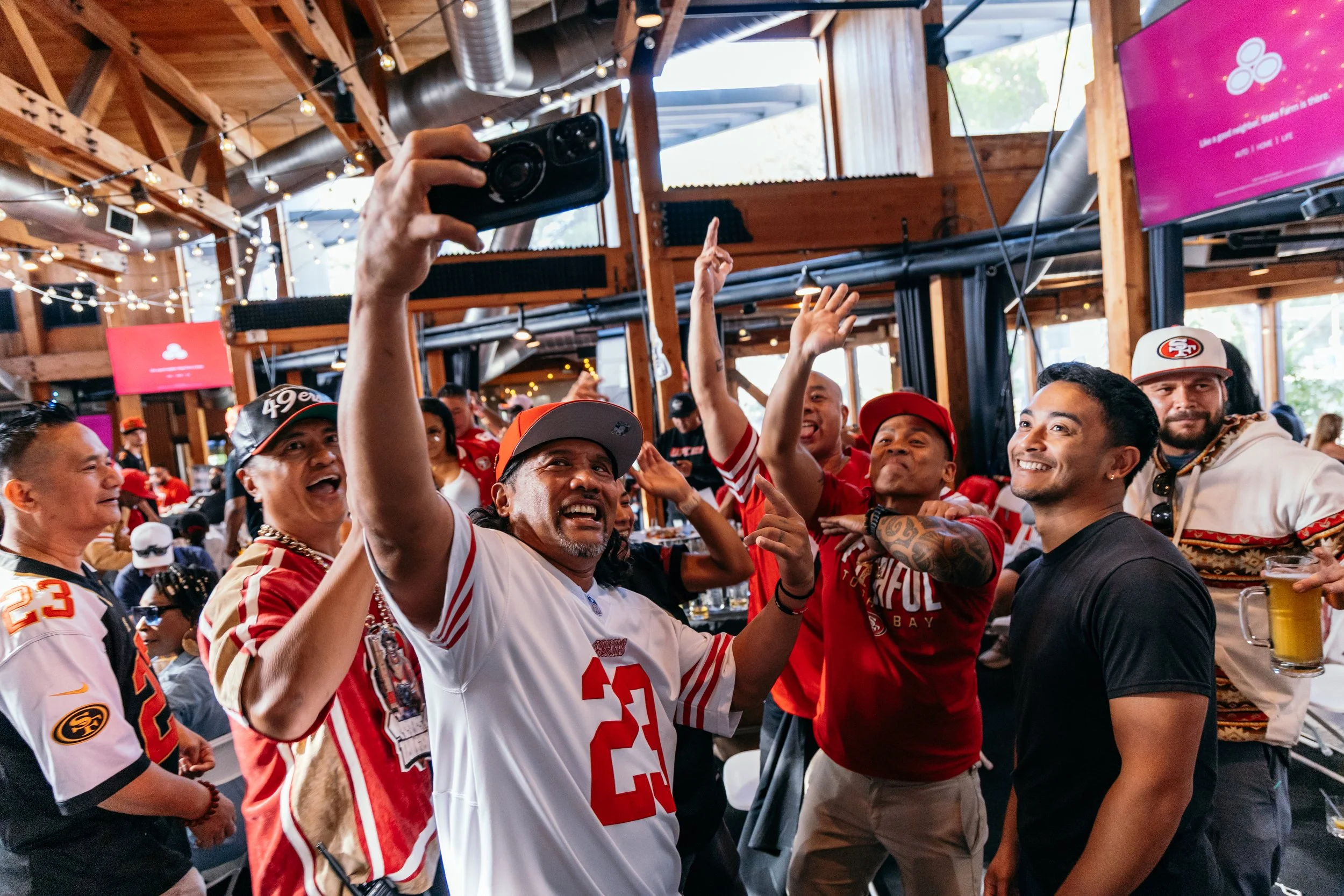 Group of grateful football fans wearing San Francisco 49ers jerseys and gear, celebrating inside a rustic bar or restaurant with wooden beams, string lights, and multiple televisions.