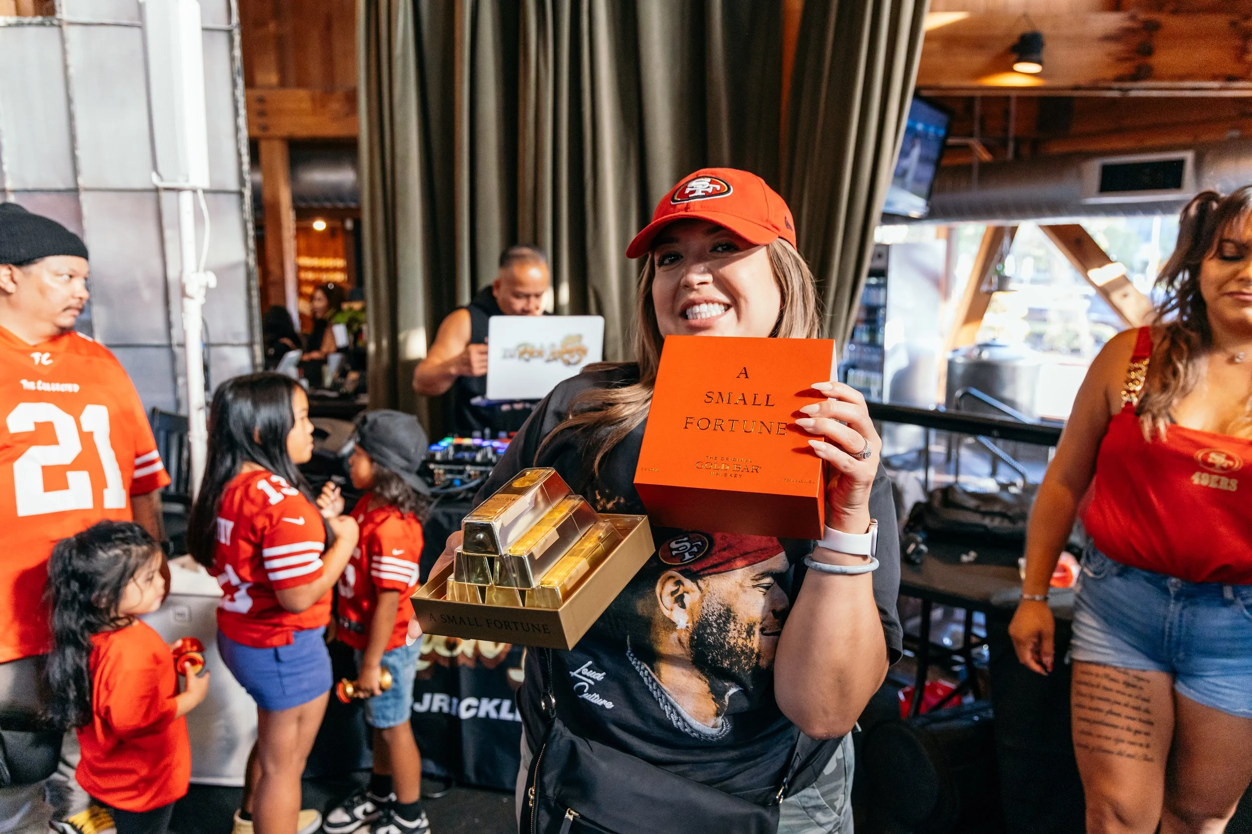 Woman in a San Francisco 49ers hat and dark jacket holding a gold bar in a crowd of people wearing red 49ers jerseys inside a wooden building.