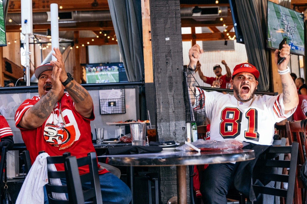 Two men wearing San Francisco 49ers jerseys cheering and celebrating at a sports bar, with multiple television screens showing a football game in the background.