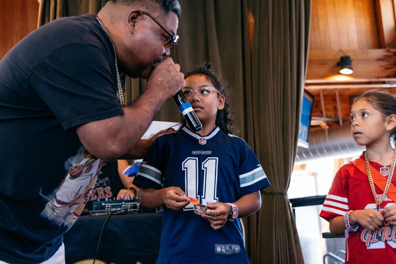 A man holding a microphone up to a young girl in a Dallas Cowboys football jersey, with another girl in a similar jersey nearby. The scene takes place indoors with wooden walls and curtains.
