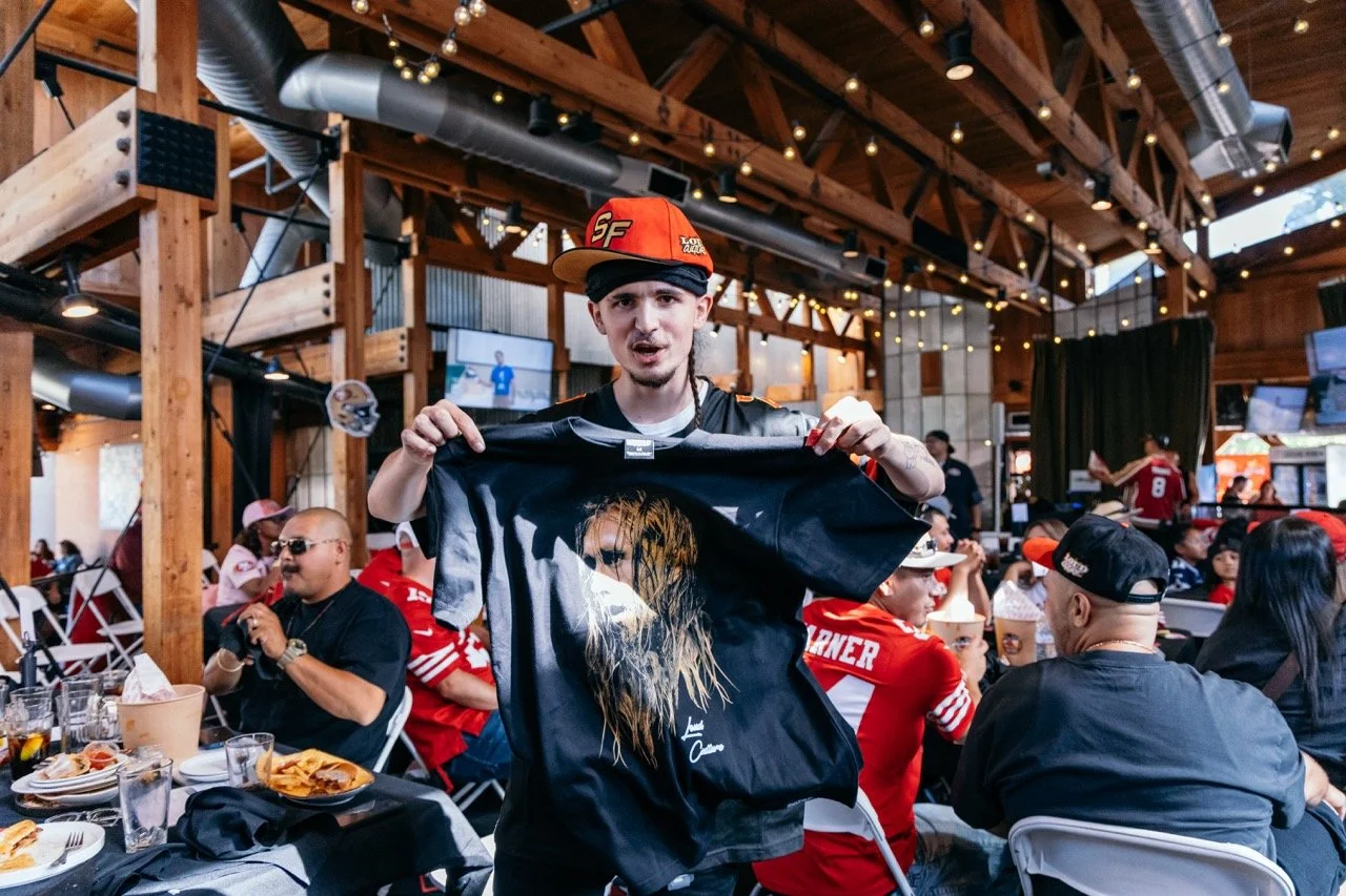 A young man in a red cap holding a black t-shirt with a woman's face printed on it at a sports bar or restaurant filled with people wearing sports jerseys