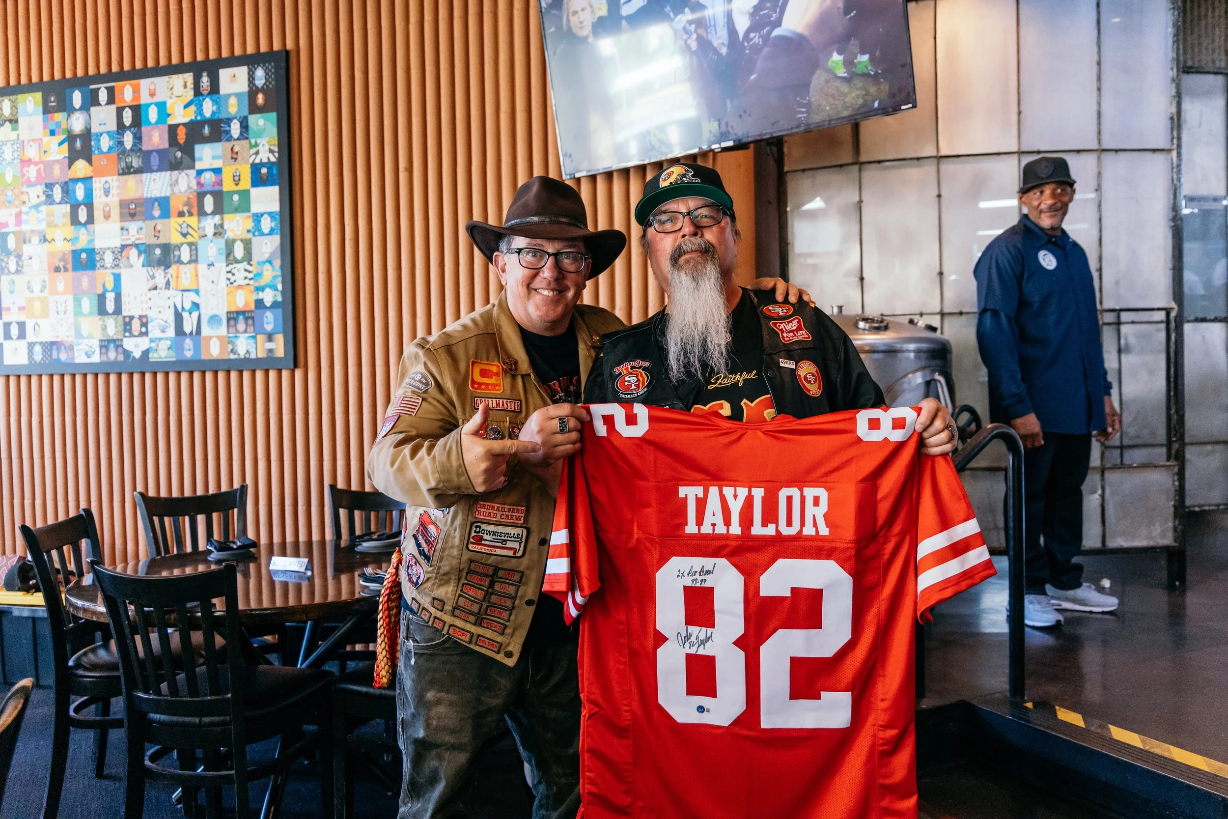 Two men are smiling and posing with a red San Francisco 49ers football jersey with the name Taylor and the number 80. One is wearing glasses, a brown cowboy hat, and a patched jacket, and the other has a long gray beard, glasses, and a cap. They are 