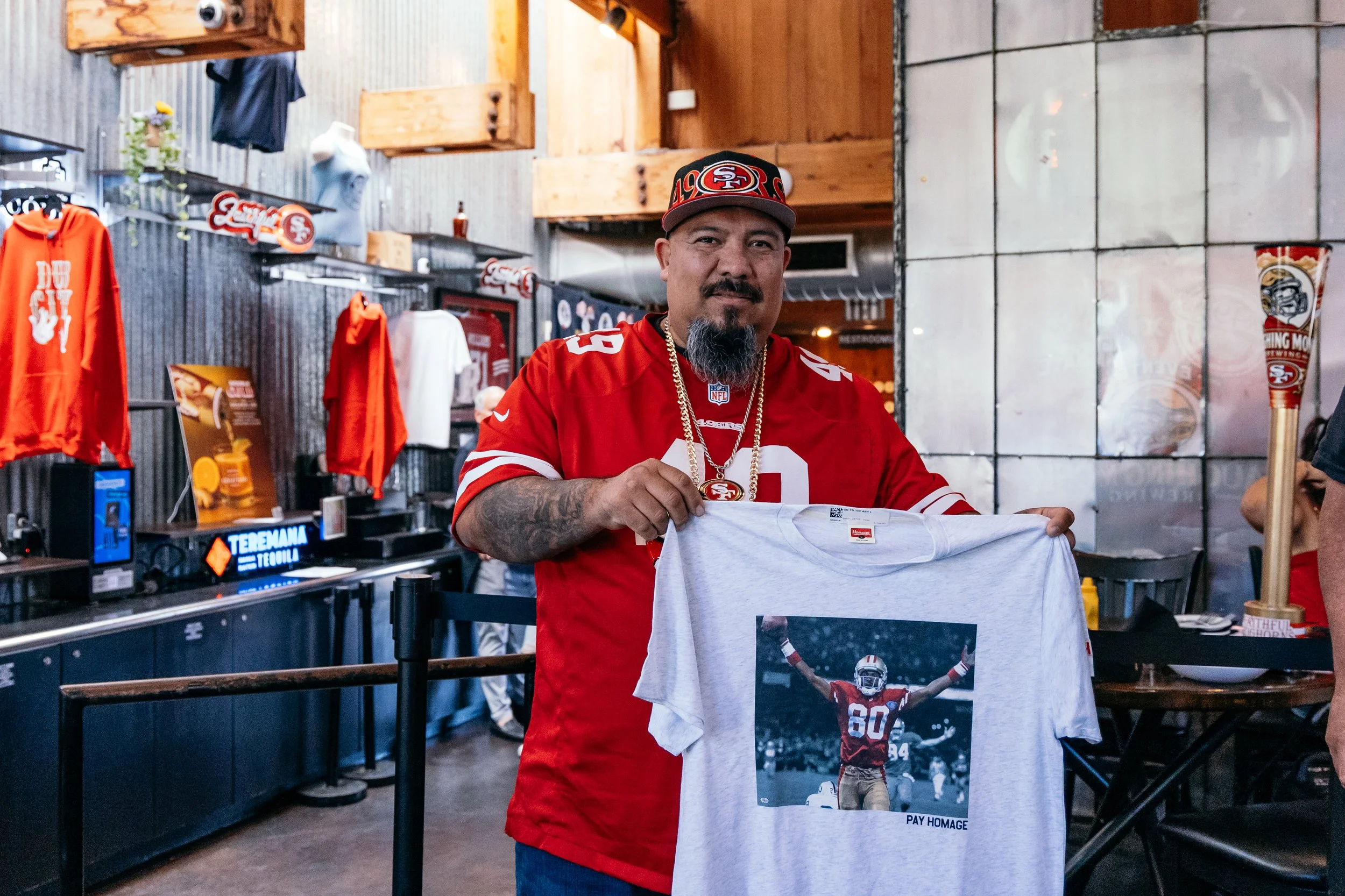A man wearing an San Francisco 49ers football jersey and a hat with the team's logo is holding a T-shirt featuring a football player in a celebratory pose. The setting appears to be a merchandise store or a fan shop, decorated with the team's colors 