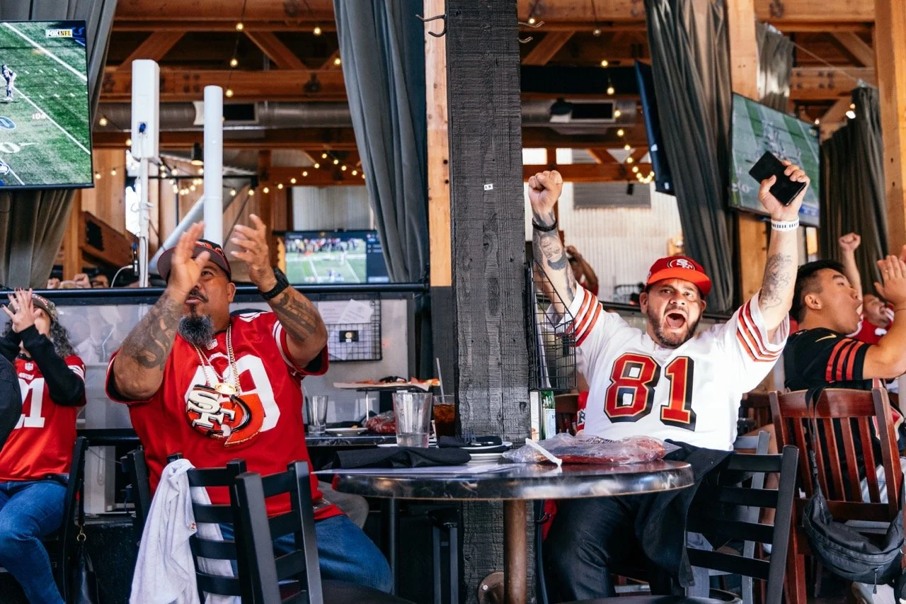 San Francisco 49ers fans cheering and celebrating during a game at a sports bar, watching multiple TV screens.