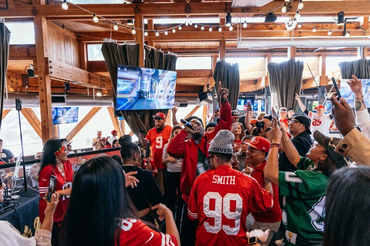 A large group of people gathered indoors, many wearing sports jerseys and hats, celebrating or at a party. Some are holding up phones, and there's a person in the center with a microphone. TV screens display a sports game or event, and the setting ha