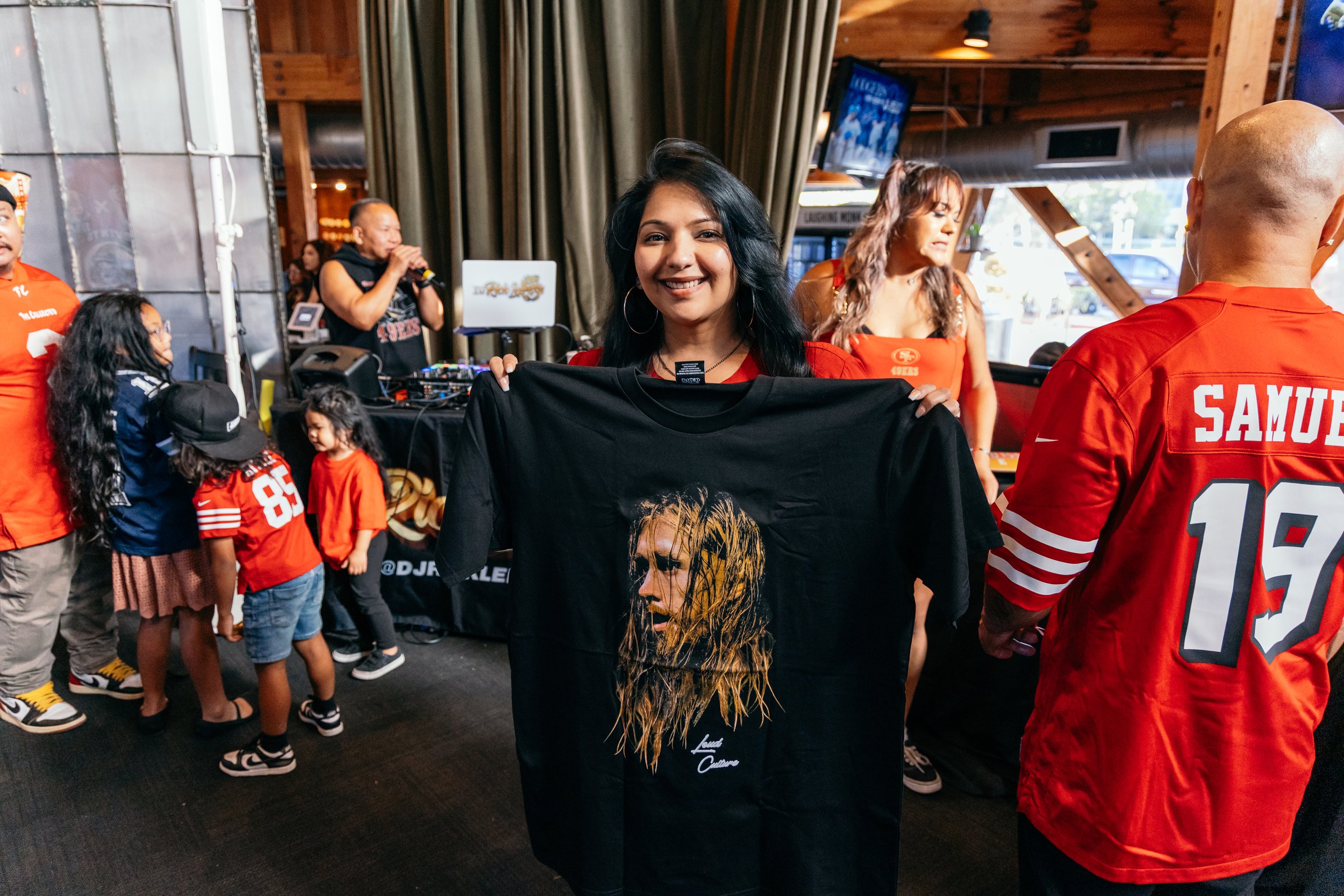 A woman with black hair holding a black t-shirt featuring a portrait, surrounded by people in sports jerseys at a sports-themed event.