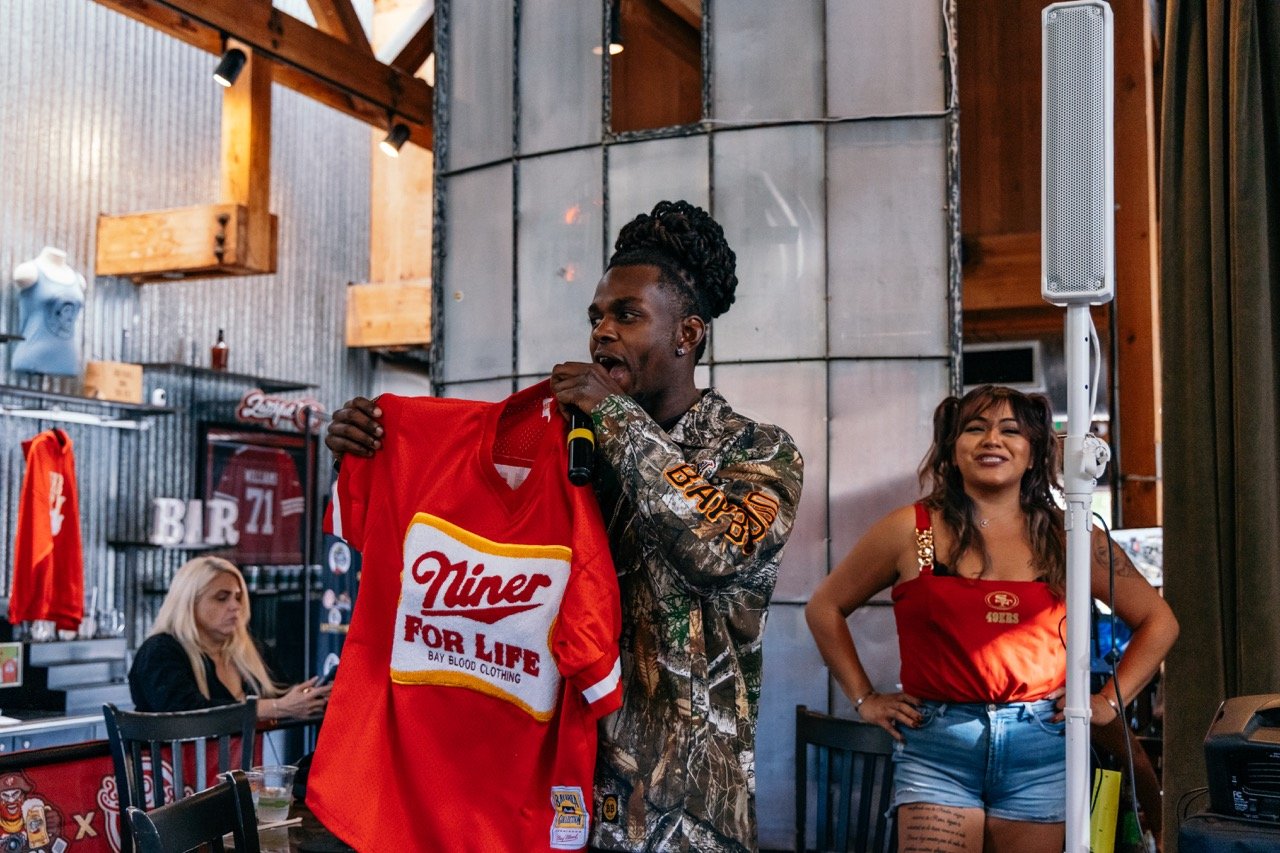 A man holds a red 'Niner for Life' shirt and is speaking into a microphone inside a bar or restaurant, with a woman standing beside him in a San Francisco 49ers tank top and denim shorts, smiling. The background shows a woman seated at a table, corru