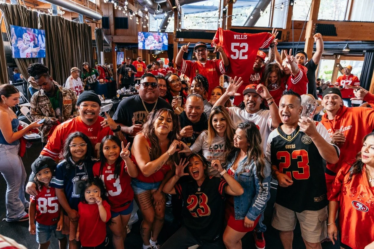 A group of San Francisco 49ers fans celebrating at a sports bar or restaurant, with some making peace signs and wearing team jerseys and shirts.