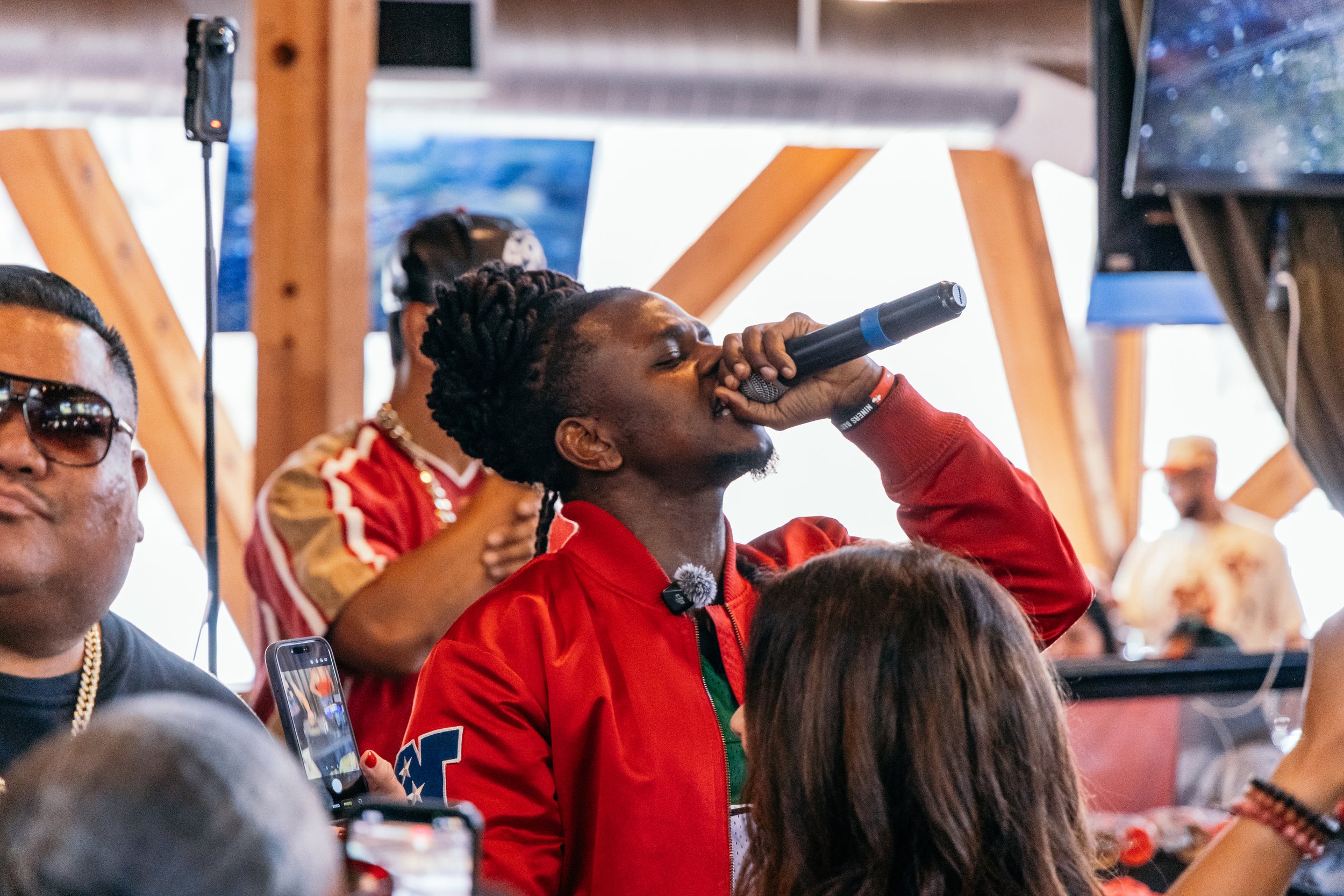 A man with dreadlocks singing into a microphone during a gathering or event with people around, some taking photos or recording.