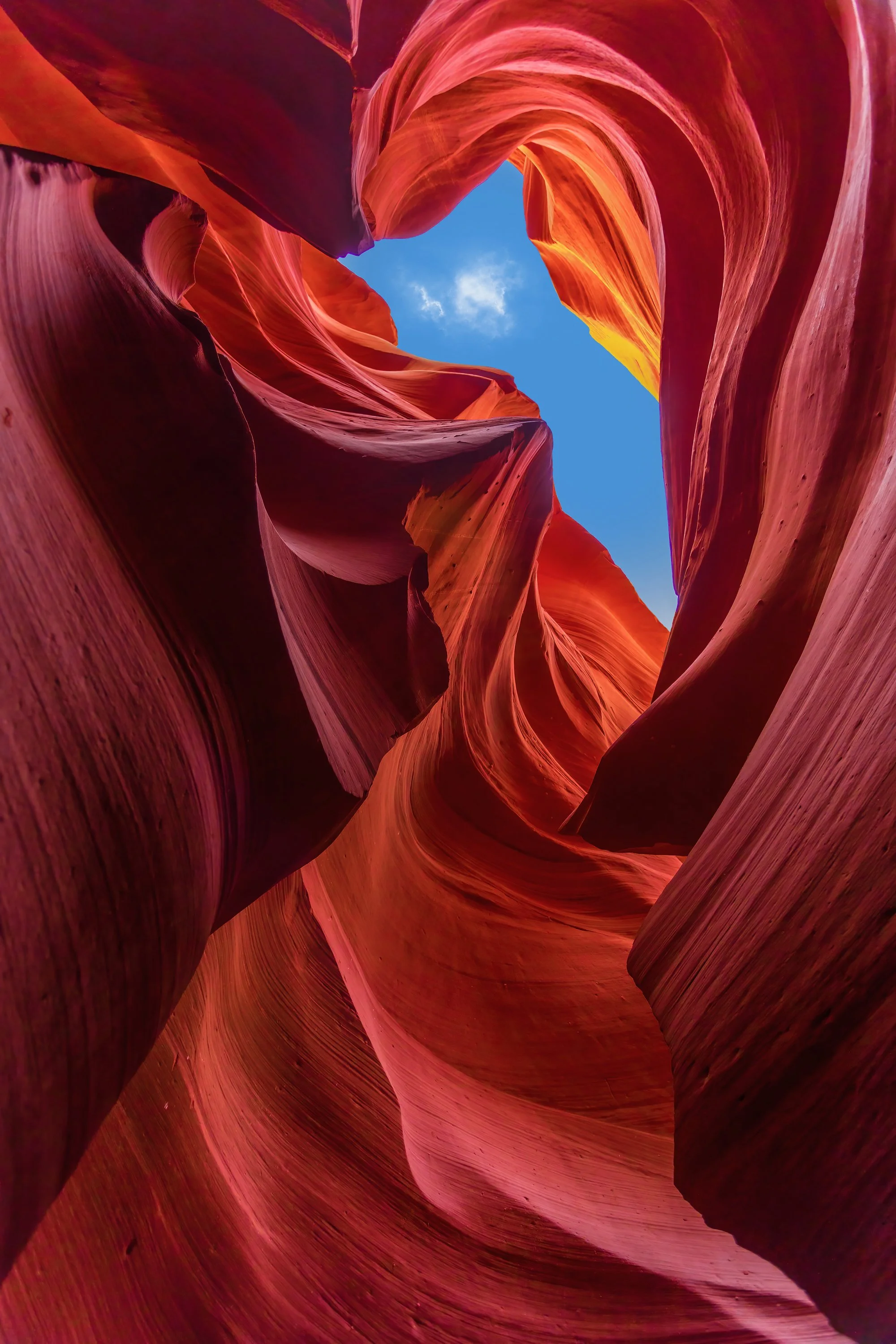 View of a narrow slot canyon with smooth, curving red and orange rock walls and a small patch of blue sky visible at the top.