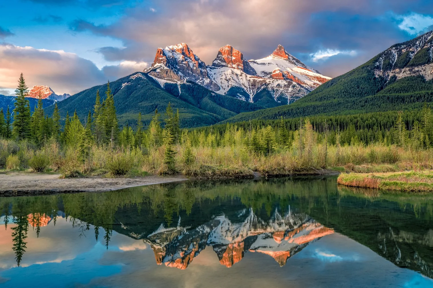 Snow-capped mountains reflecting in a calm river, surrounded by green trees and a partly cloudy sky.