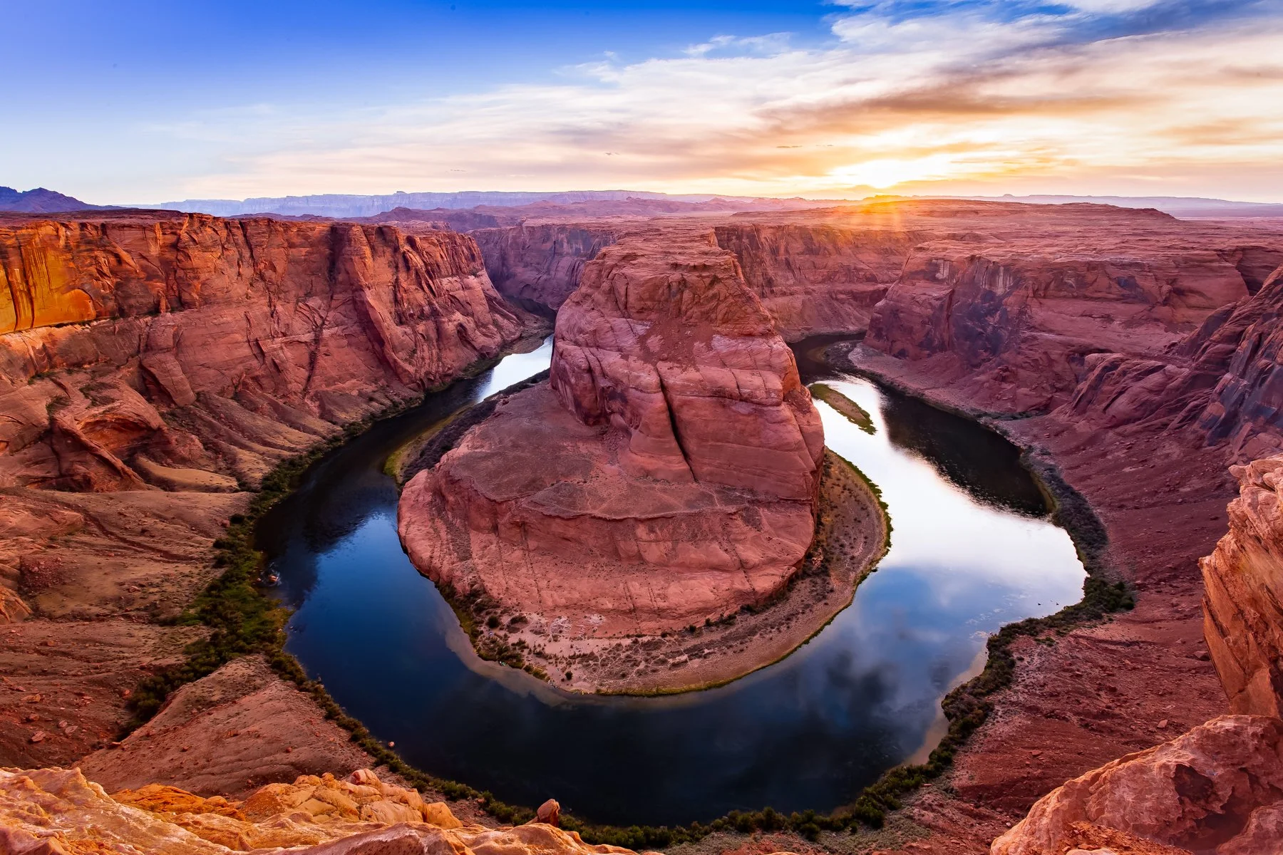 Arizona's Horseshoe Bend at sunset, with a meandering river forming a dramatic horseshoe shape around a large central rock formation, red canyon walls, and a colorful sky with clouds and the setting sun.