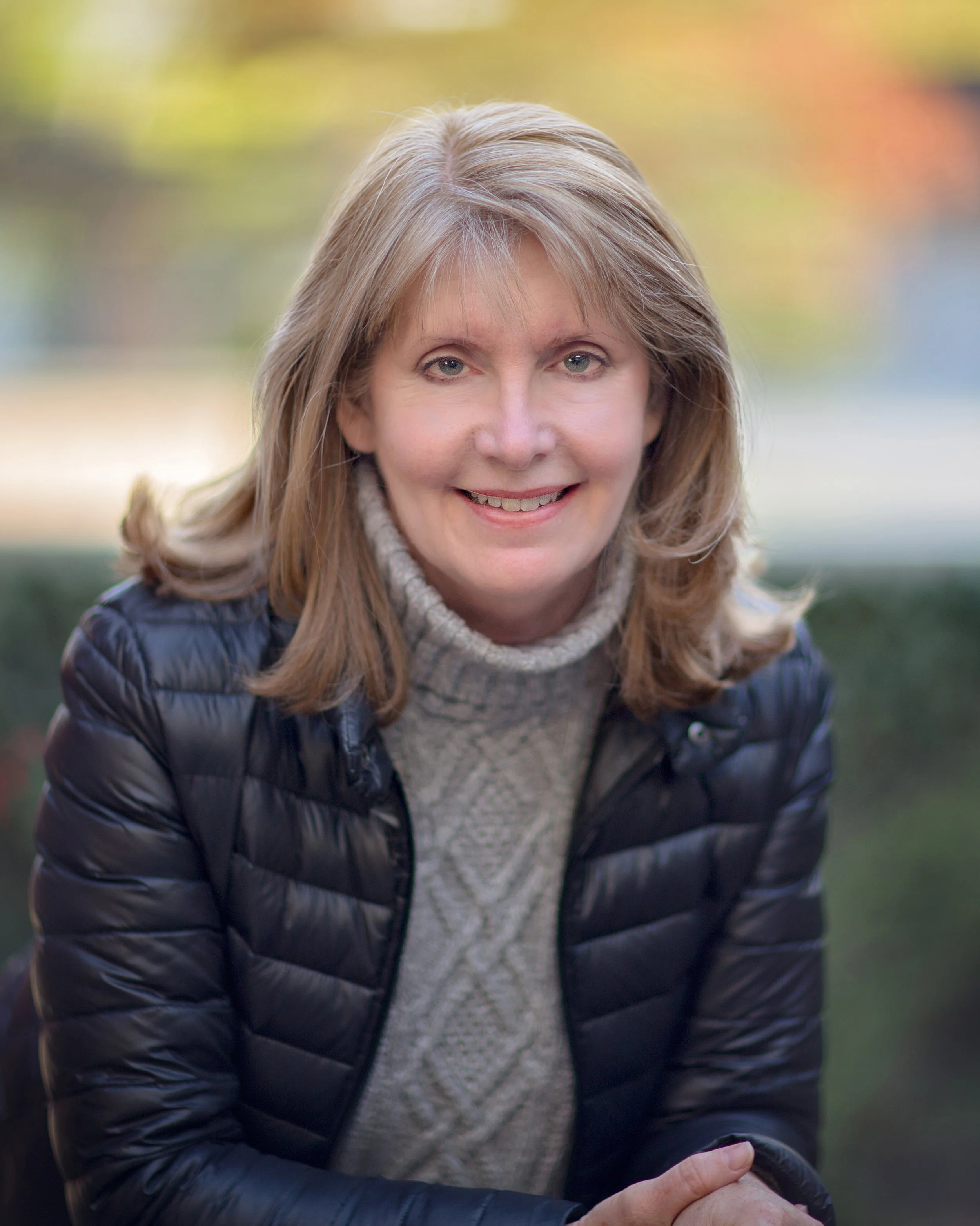 A middle-aged woman with shoulder-length blonde hair, wearing a gray turtleneck sweater and a black quilted jacket, smiling outdoors with a blurred colorful background.