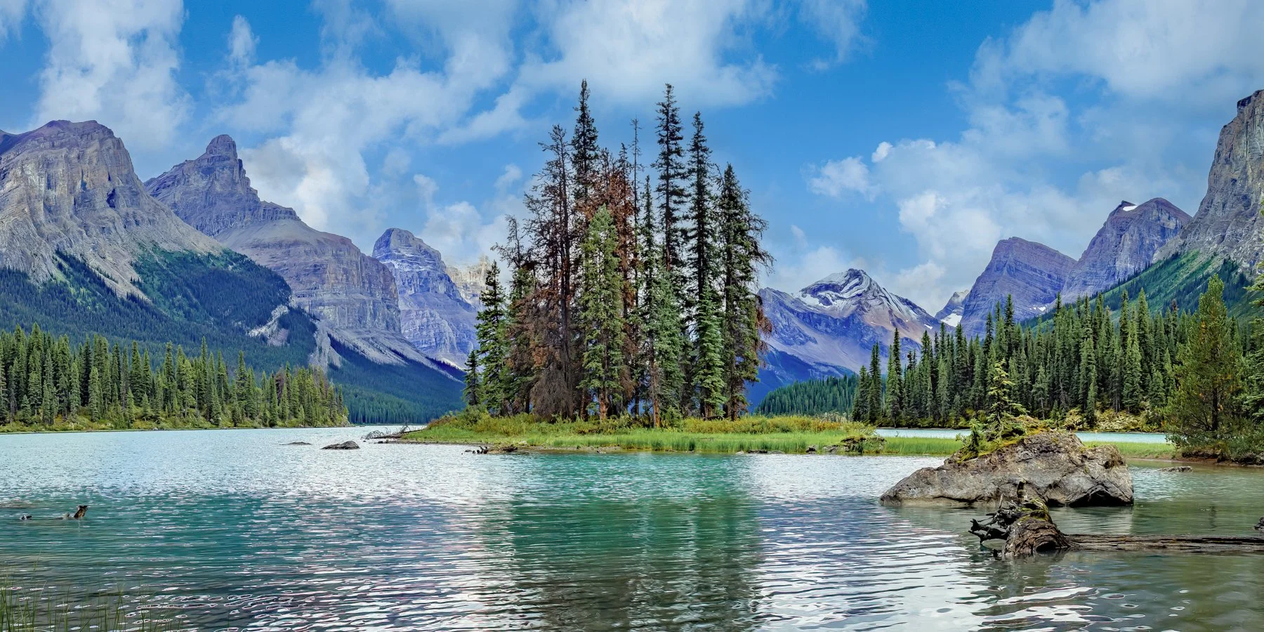 A scenic view of a serene lake with a small island covered in tall evergreen trees, surrounded by forested mountains under partly cloudy sky.