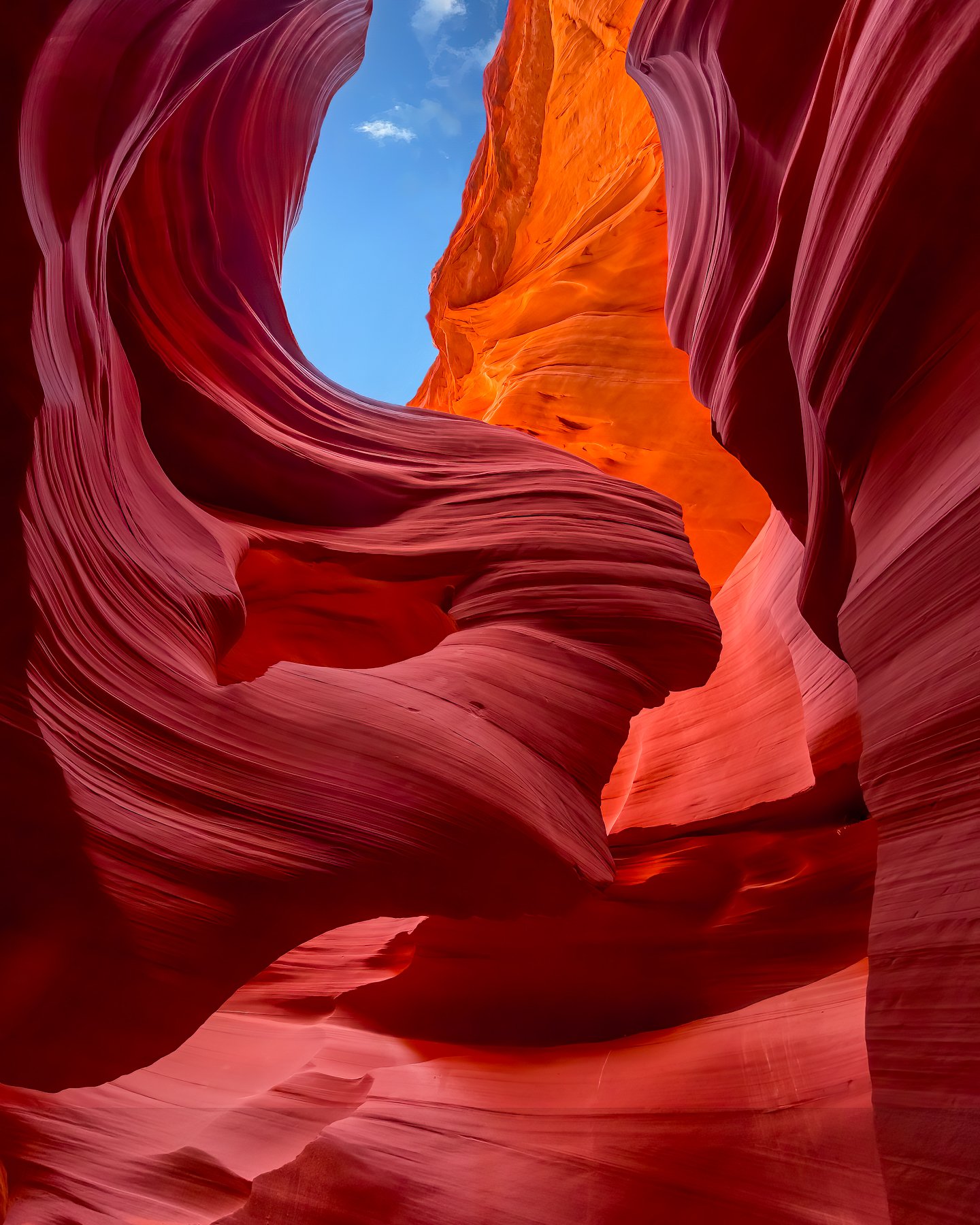 View of a narrow slot canyon with red and orange layered rock formations, a small patch of blue sky and a few clouds visible at the top.