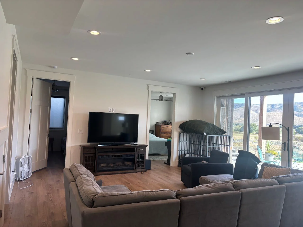 Living room with beige sofa, black armchair, TV on stand, sliding glass door showing outdoor landscape with mountains, ceiling lights, and a view into an adjacent bedroom.