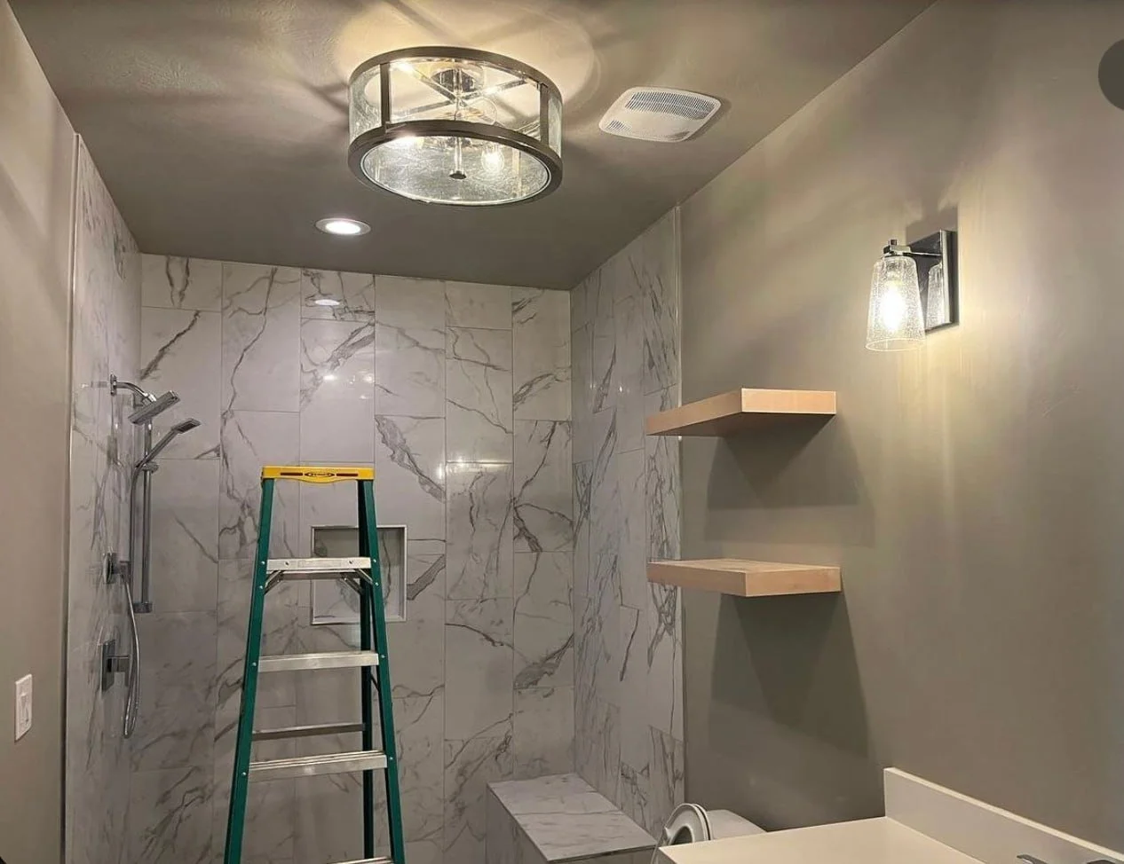 Bathroom under renovation with marble tiled shower area, ladder, wall-mounted shelves, and wall-mounted light fixture.