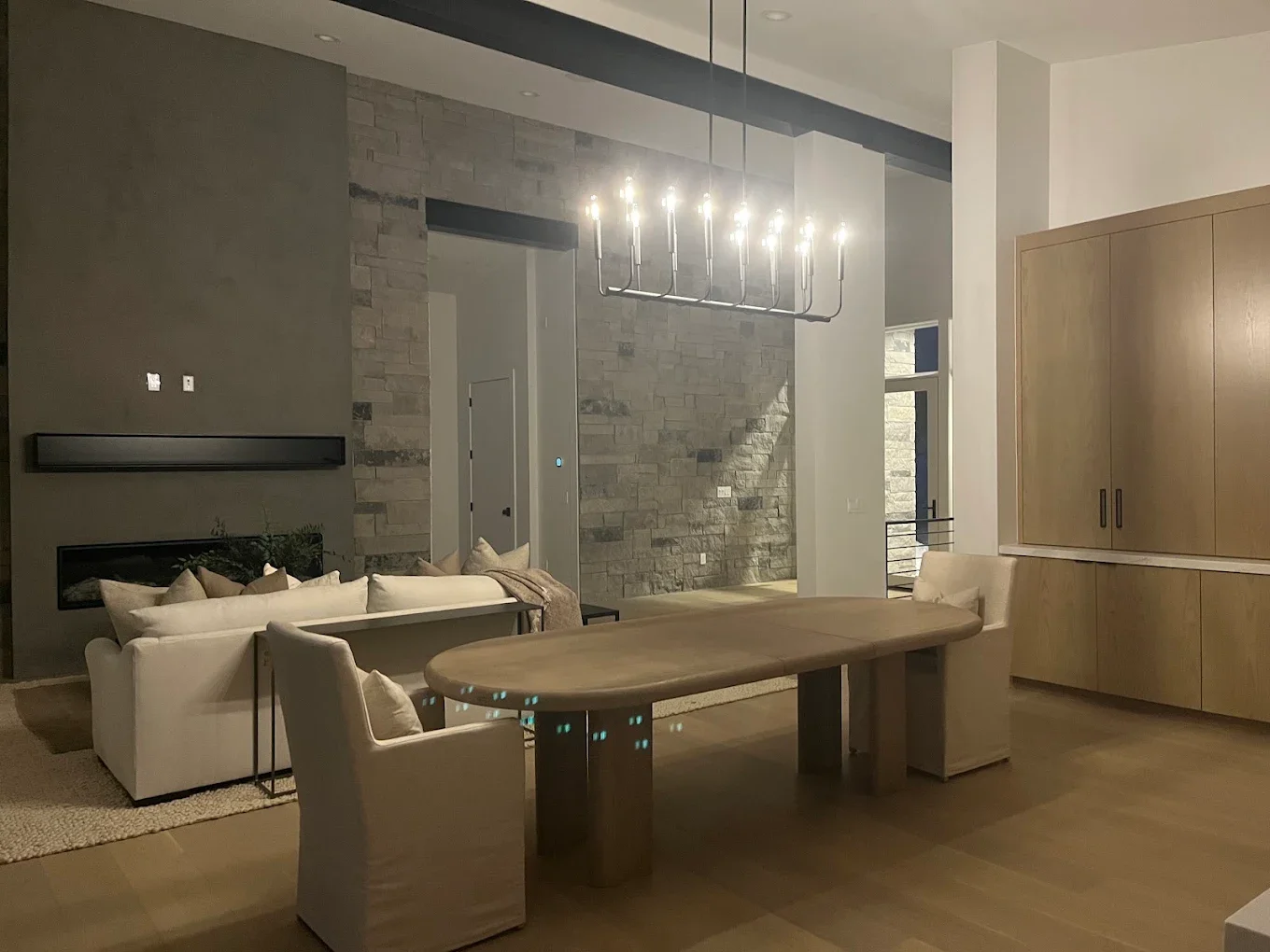 Modern living room with a wooden dining table, beige chairs, a white sofa with beige cushions, a stone accent wall, a chandelier, and light wood cabinets.