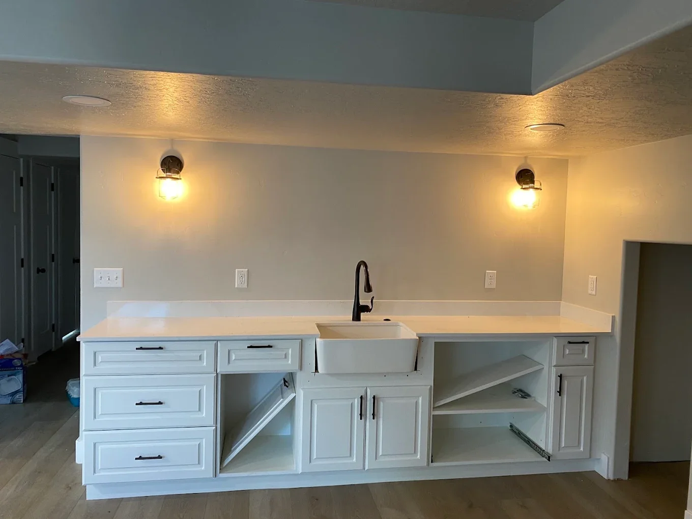 Empty kitchen with white cabinets, a farmhouse sink, and black hardware, with unfinished drawers and open shelving, illuminated by two wall-mounted lights.