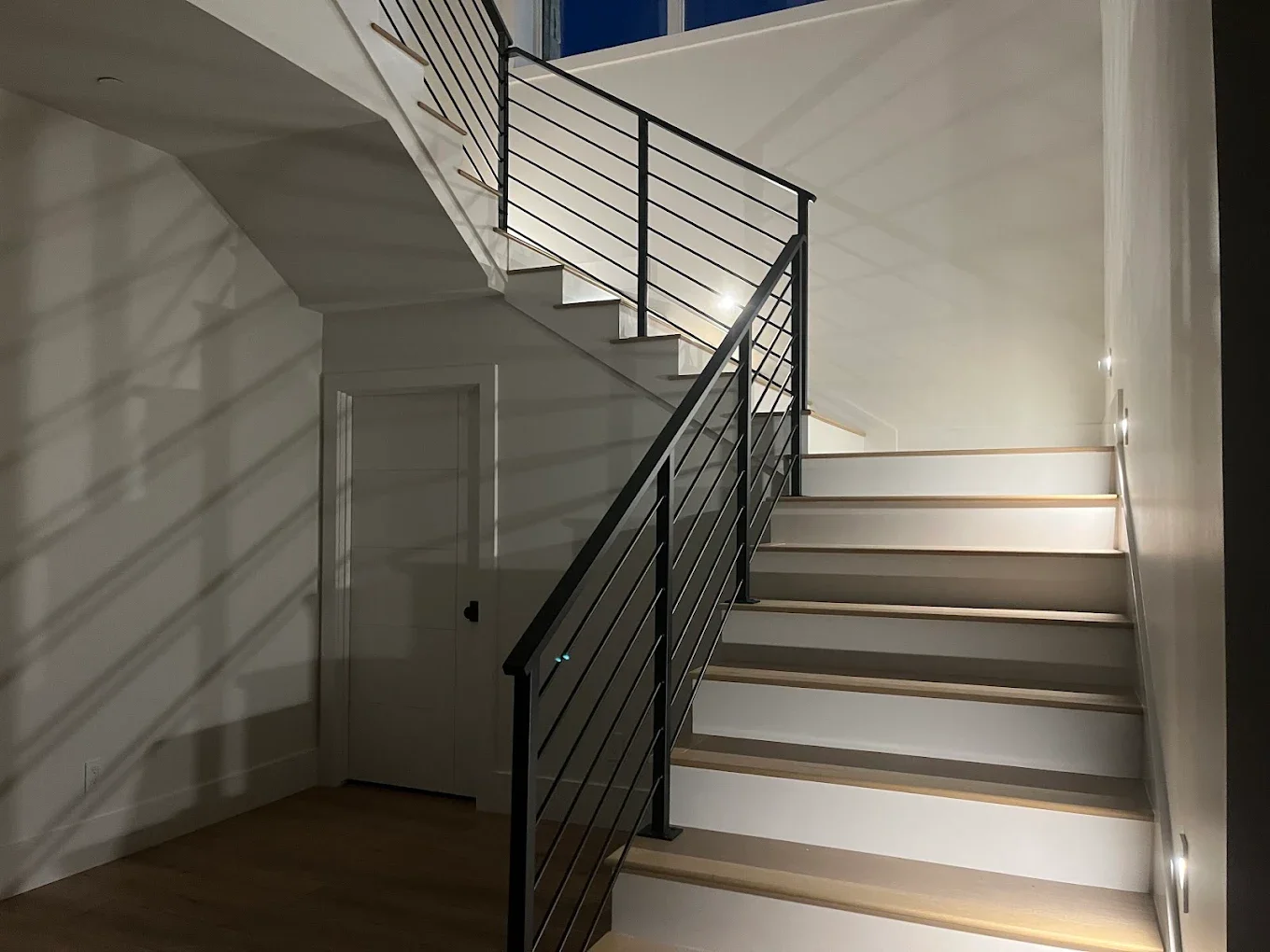 Modern staircase with black metal railings and white steps, illuminated by small wall lights, in a minimalist interior.
