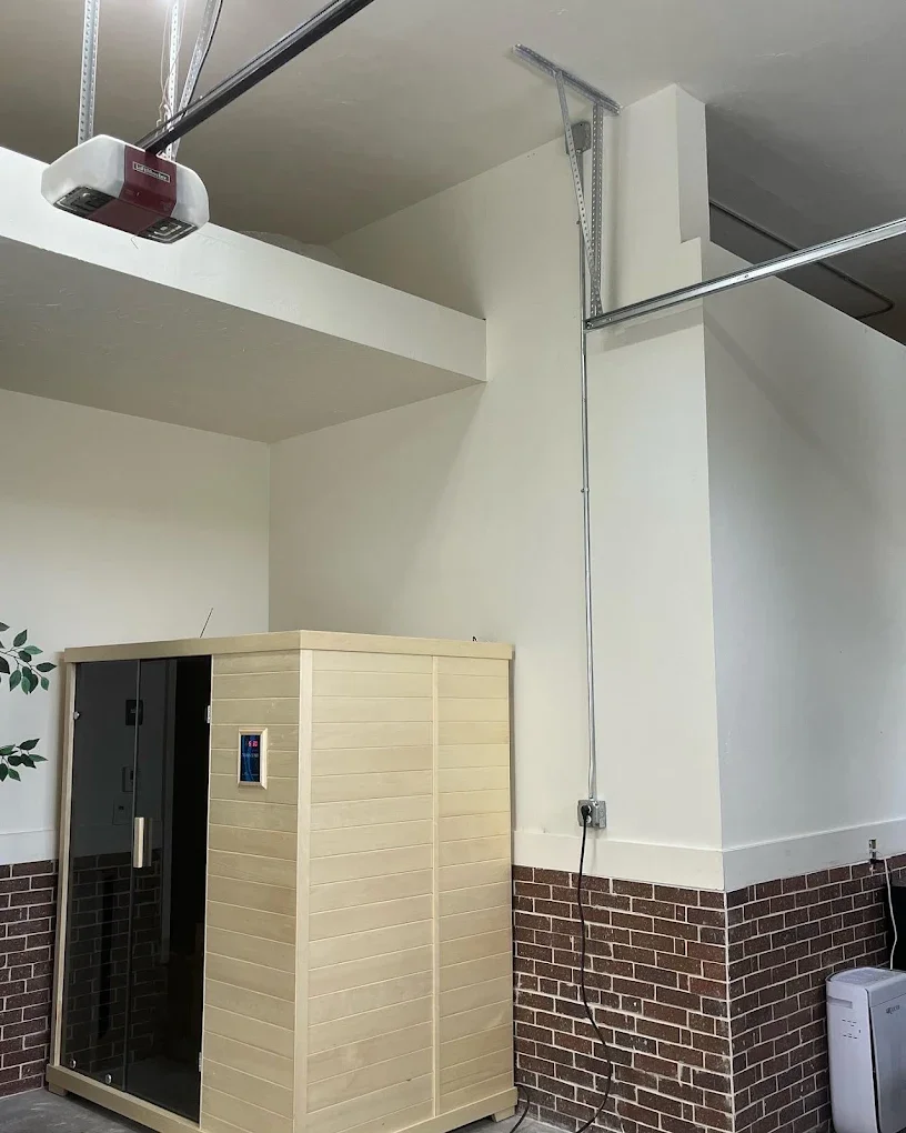 An indoor space with a small wooden sauna and a sauna thermometer on the door. A garage door opener is mounted on the ceiling, and metal tracks for a roll-up garage door are visible. An electrical outlet with a cord plugged in is on the wall near the