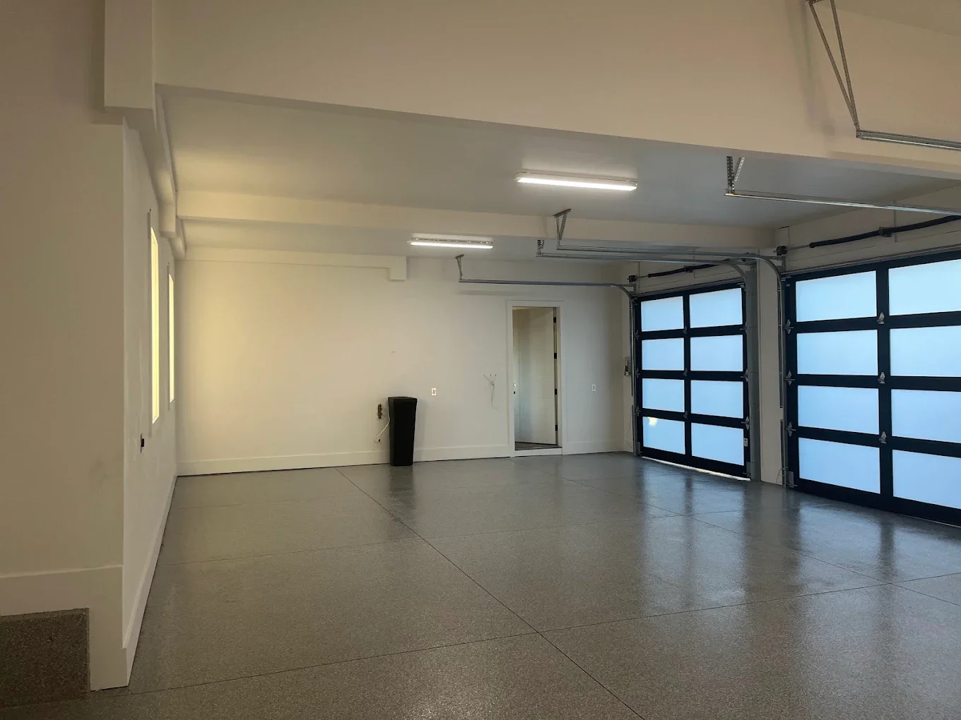Empty garage with a speckled gray floor, white walls, two garage doors with blue panels, and fluorescent ceiling lights.