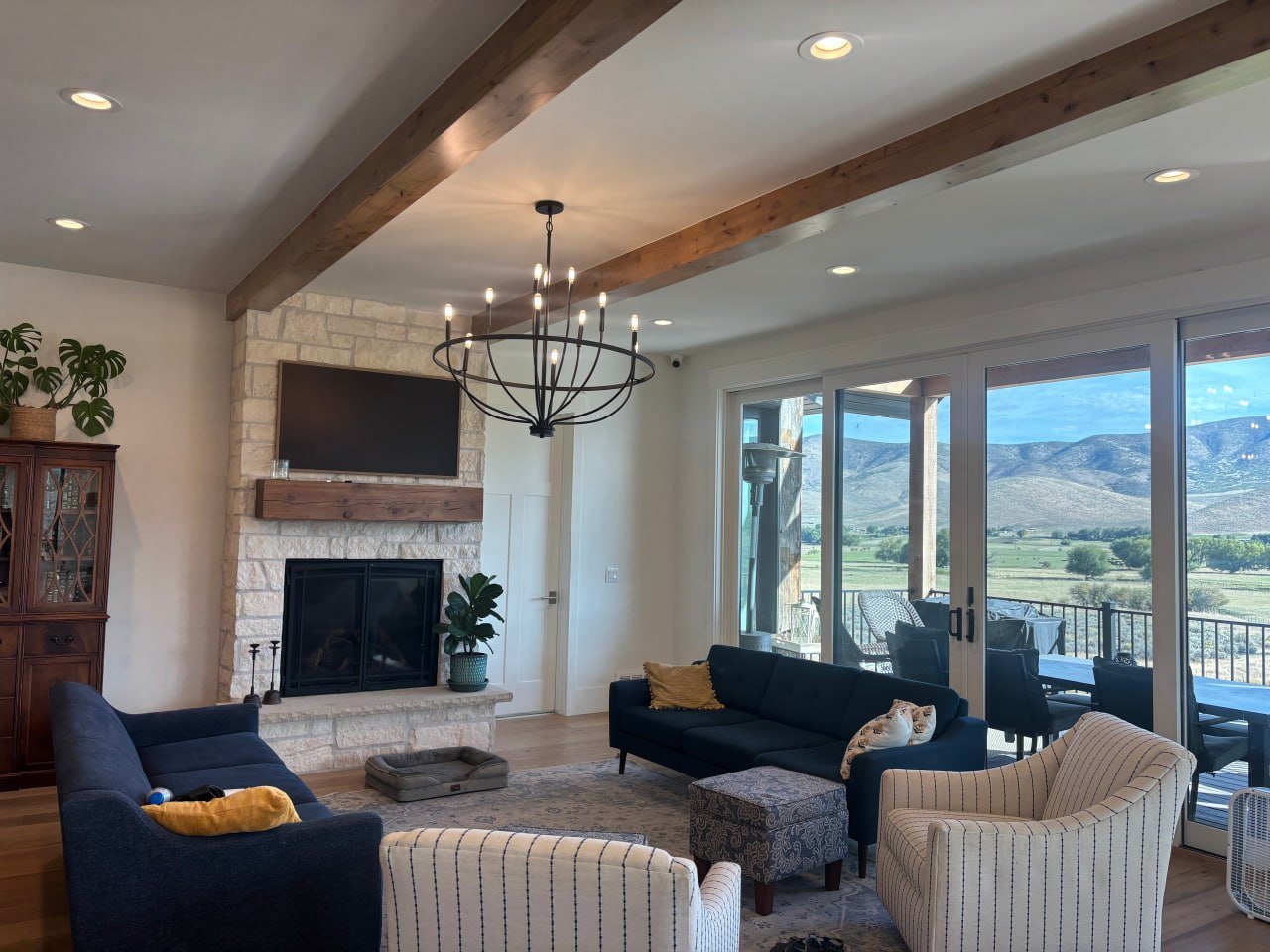 Living room with stone fireplace, flat-screen TV, wooden mantel, navy blue and white striped armchairs, a dark blue sofa, decorative pillows, a chandelier, wood beams on ceiling, large sliding glass doors leading to a patio with mountain views.