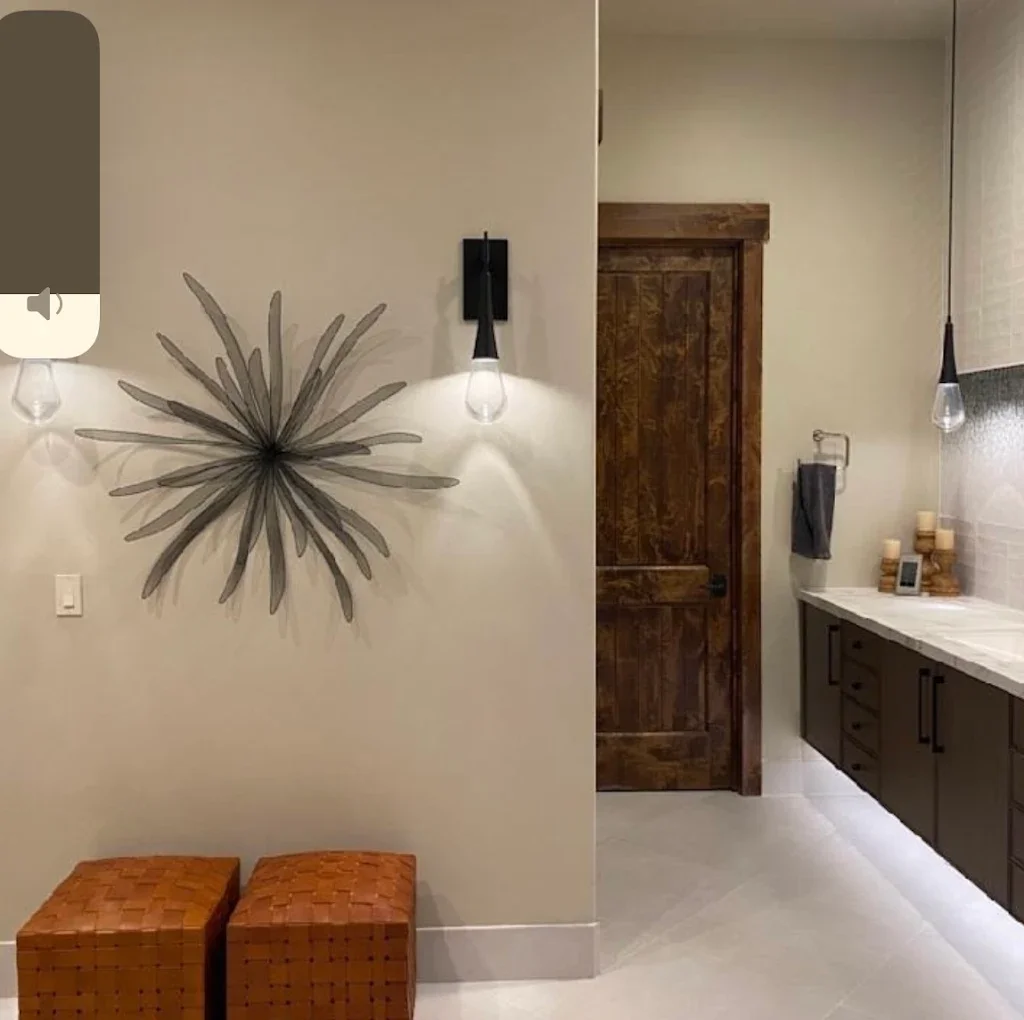 Interior view of a modern room with two orange woven stools, wall-mounted lights, a decorative wall sculpture, a wooden door, and a bathroom vanity with a mirror and candles.