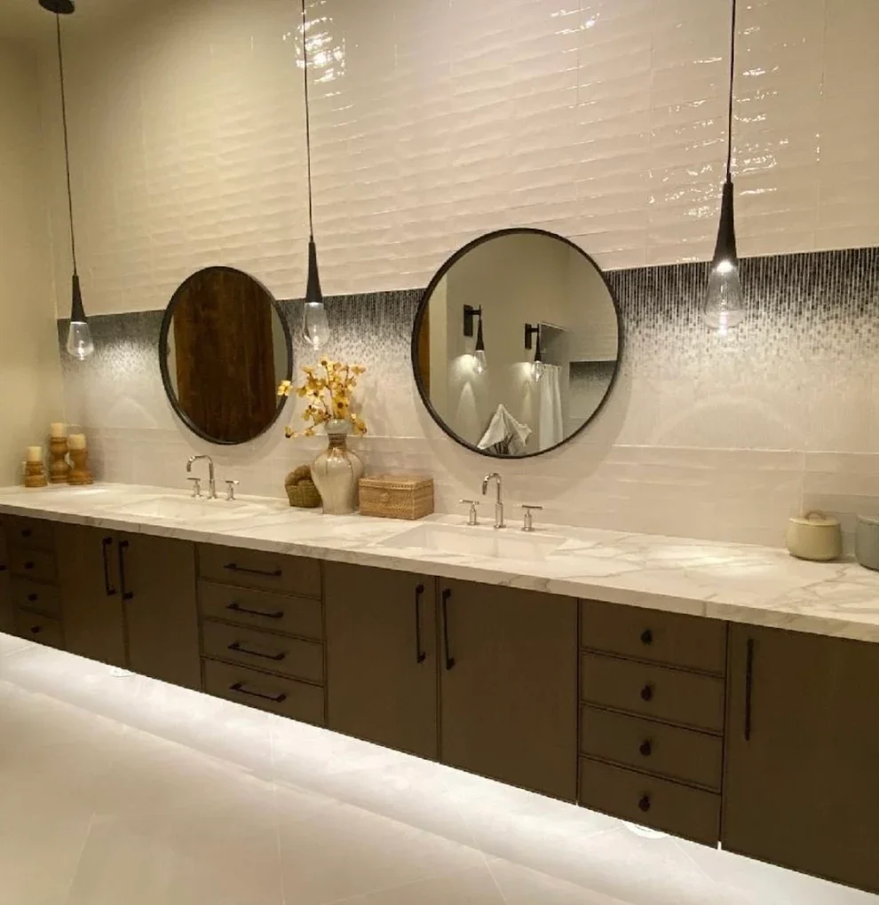 Modern bathroom with a long marble countertop, two round mirrors, pendant lights, potted plant, and bathroom accessories.