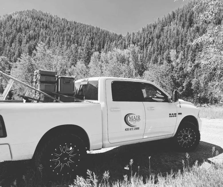 A white pickup truck parked on grass with trees and mountains in the background, carrying boxes and tools in the truck bed.