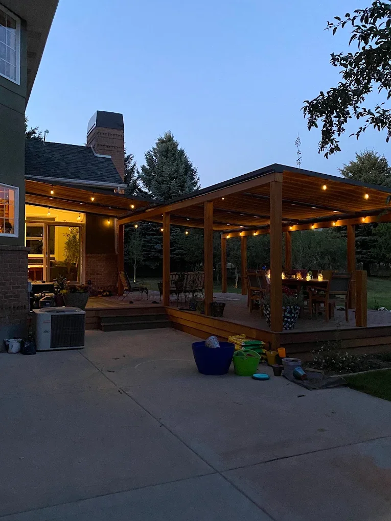 Backyard patio with a wooden deck and a covered outdoor dining area decorated with string lights, at dusk. There are toys and buckets on the concrete patio, and a house with a lit interior visible through a sliding glass door.