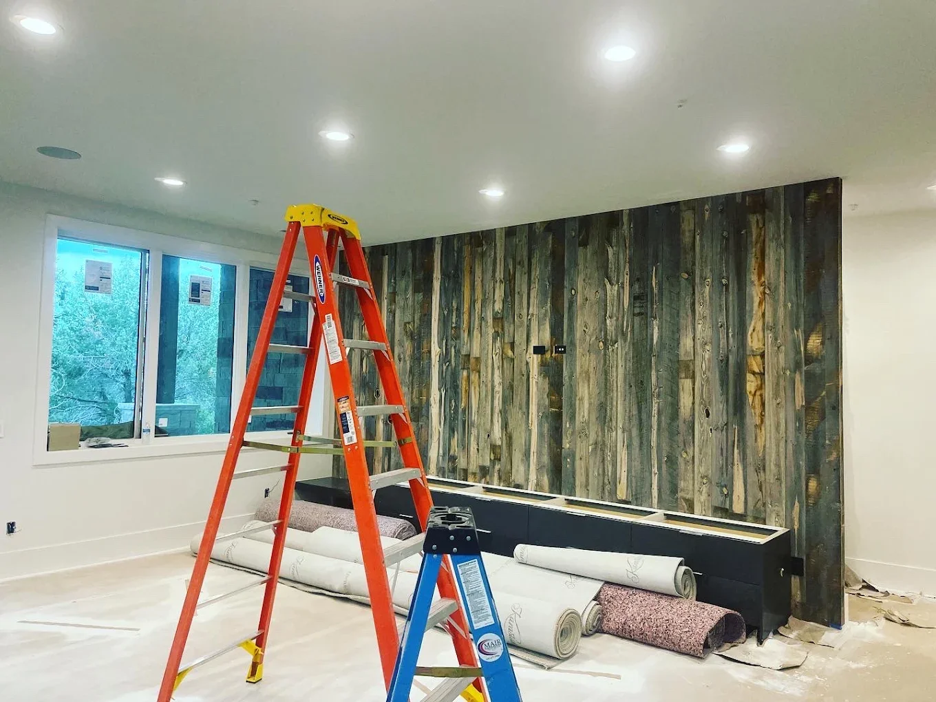 Living room under renovation with a ladder, unfinished wall paneling, and construction materials on the floor.