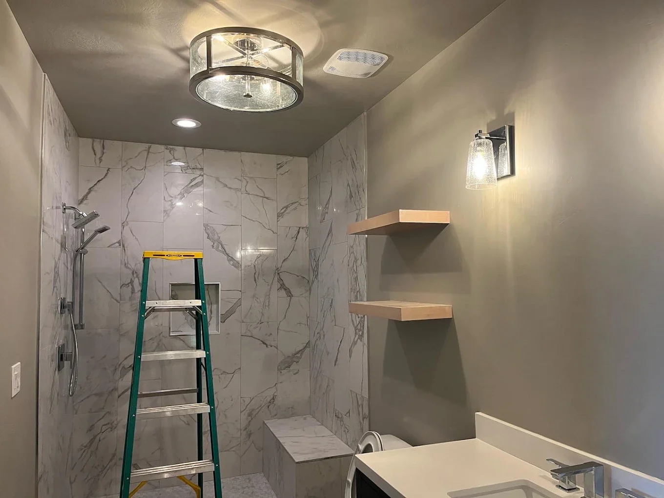 Bathroom under renovation with a ladder, marble-tiled shower area, wall mounted shelves, ceiling light fixture, and partially installed sink.