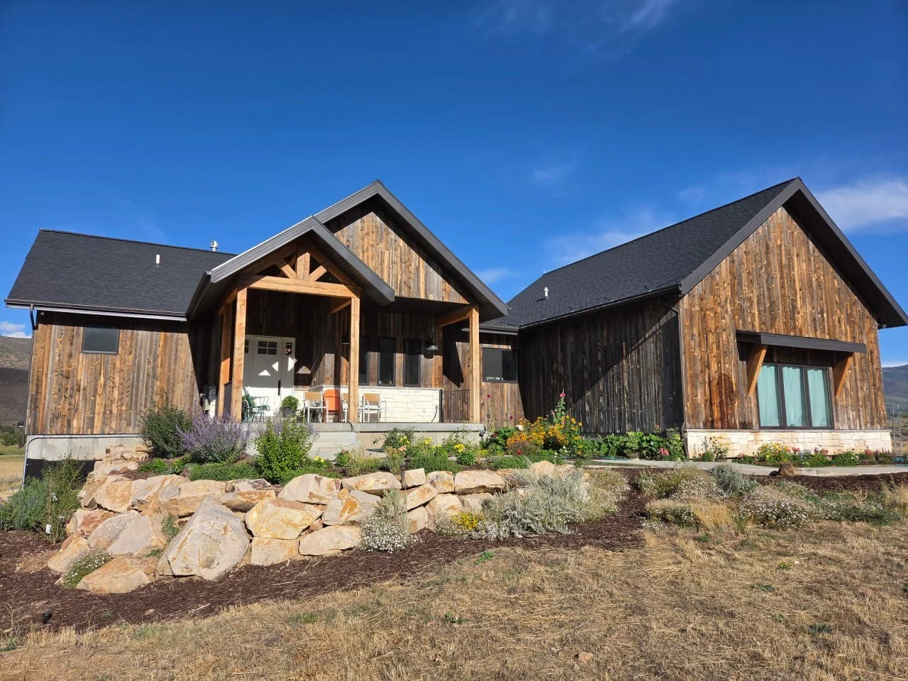 A modern house with wooden siding, black roof, and a front porch, surrounded by a landscaped garden with rocks and flowers under a clear blue sky.