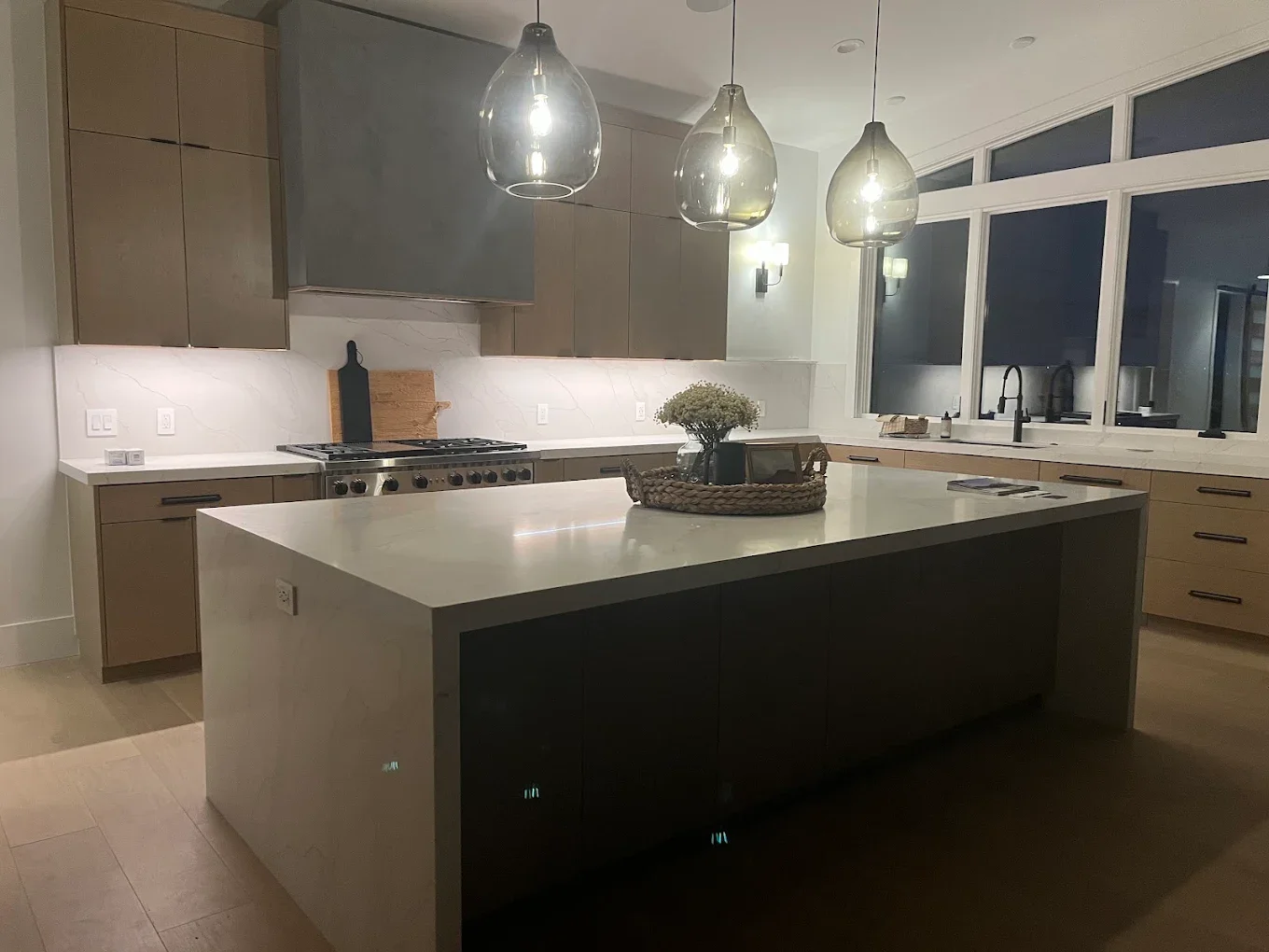 Modern kitchen with a central island, three pendant lights above, beige cabinets, a white marble backsplash, and large windows.