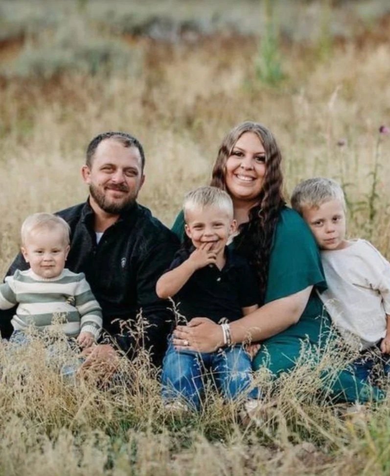 A family of five sitting outdoors in a field of tall grass, smiling at the camera. The family includes two adults, a man and a woman, and three young children.