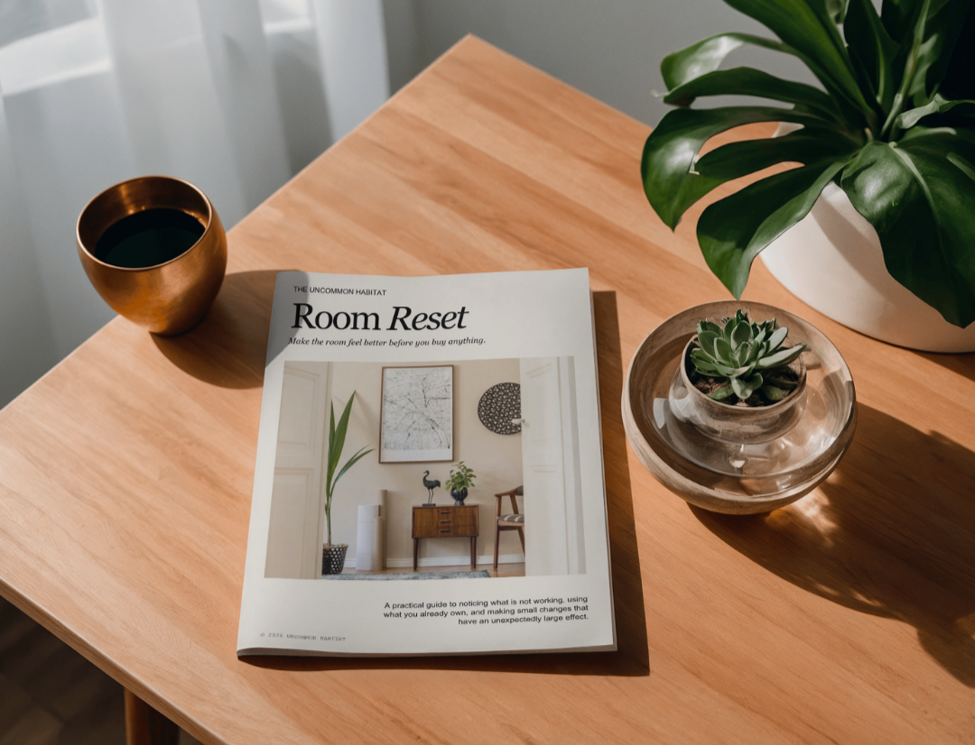 A wooden table with a magazine titled 'Room Reset,' a cup of black coffee in a copper mug, and two potted plants, one in a transparent glass bowl and the other in a white pot, with natural light illuminating the scene.
