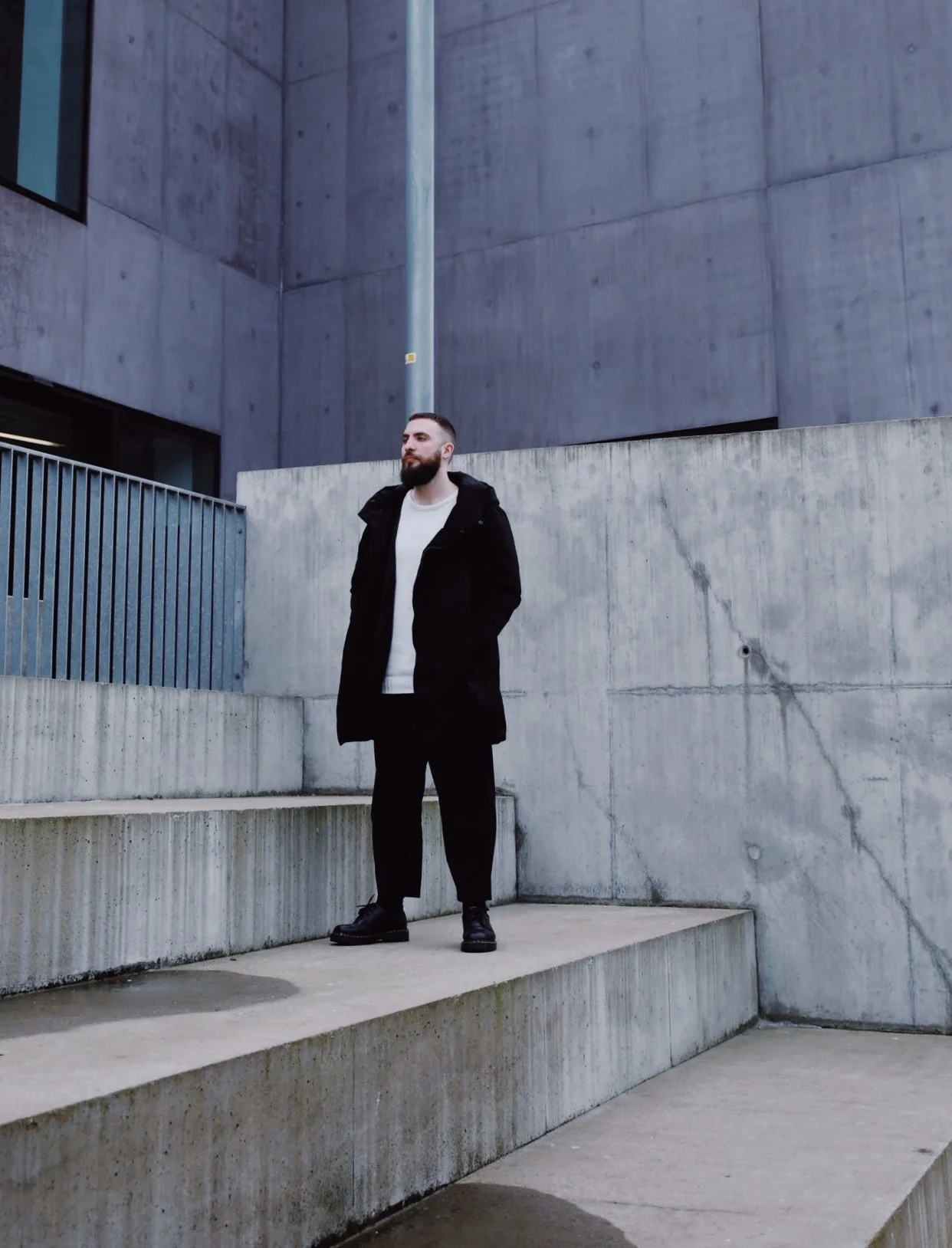 A man with a beard dressed in a black coat, black pants, and black shoes stands on concrete steps outside against a large concrete building with minimal architectural details.