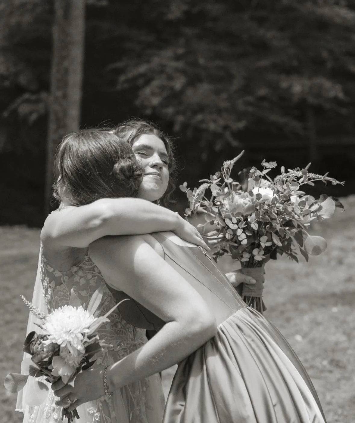 Two women hugging, one holding a bouquet of flowers, in an outdoor setting.