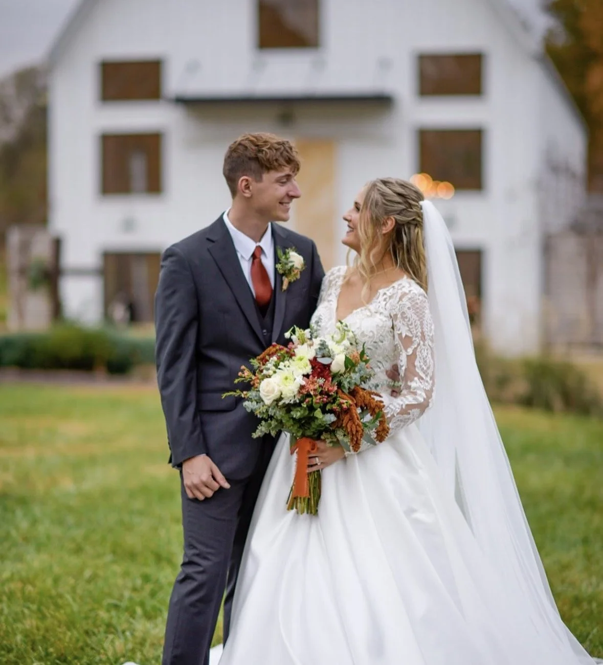 A bride and groom in wedding attire stand outdoors, smiling and looking at each other. The bride is holding a bouquet of colorful flowers. A white building is in the background.